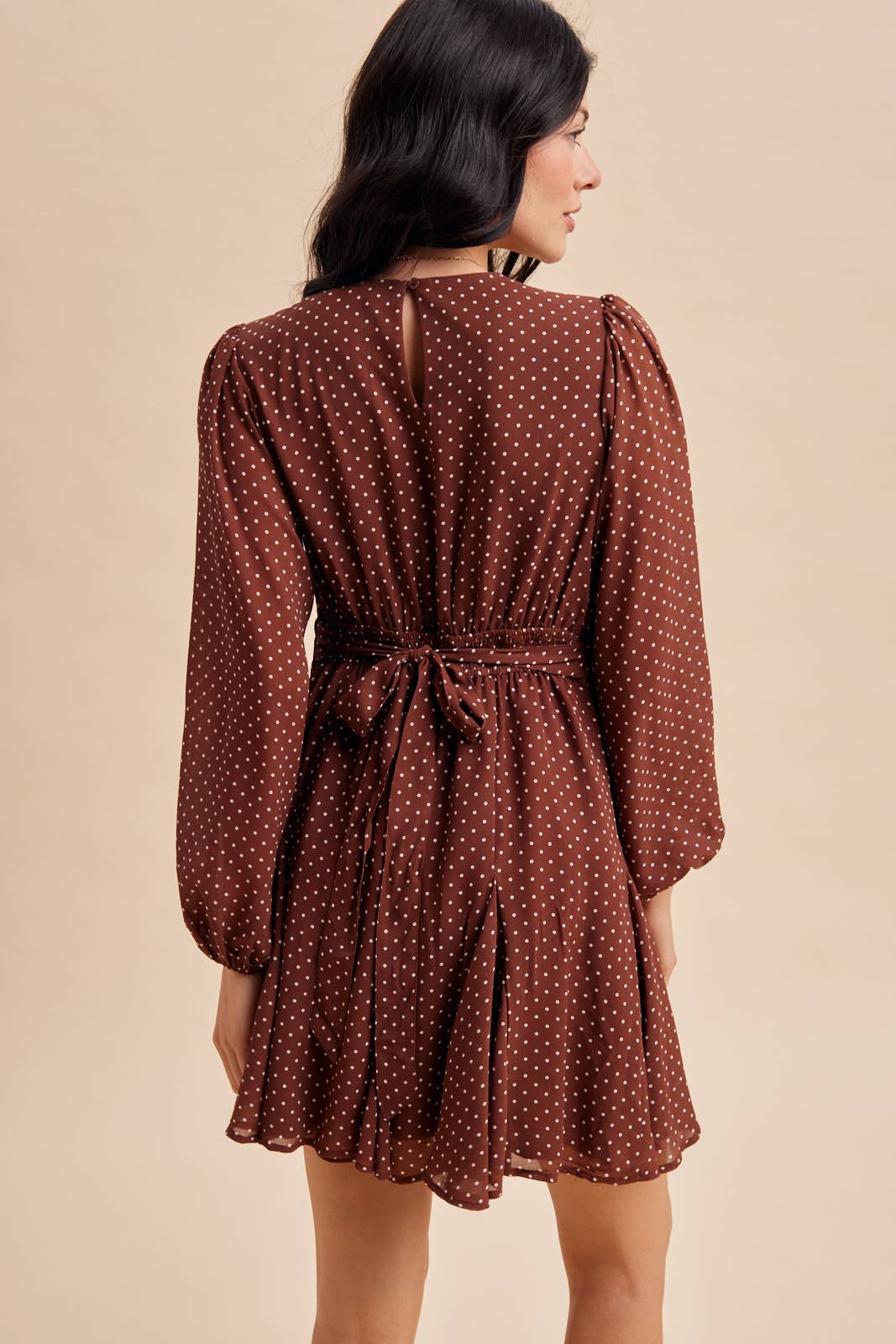 A woman viewed from behind wearing an In Loom Polka Dot Flared Mini Dress, featuring a brown long-sleeved design with a white polka dot pattern and a tie-waist.