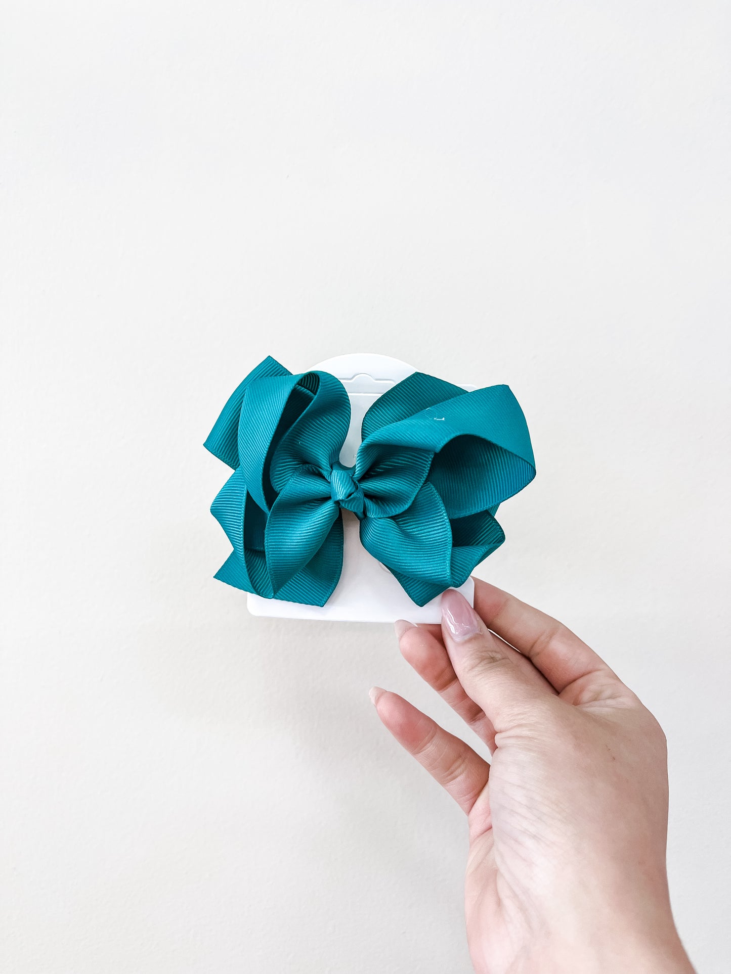 A hand holds a funteze Classic Boutique Bow in teal, attached to a white card against a plain light background.