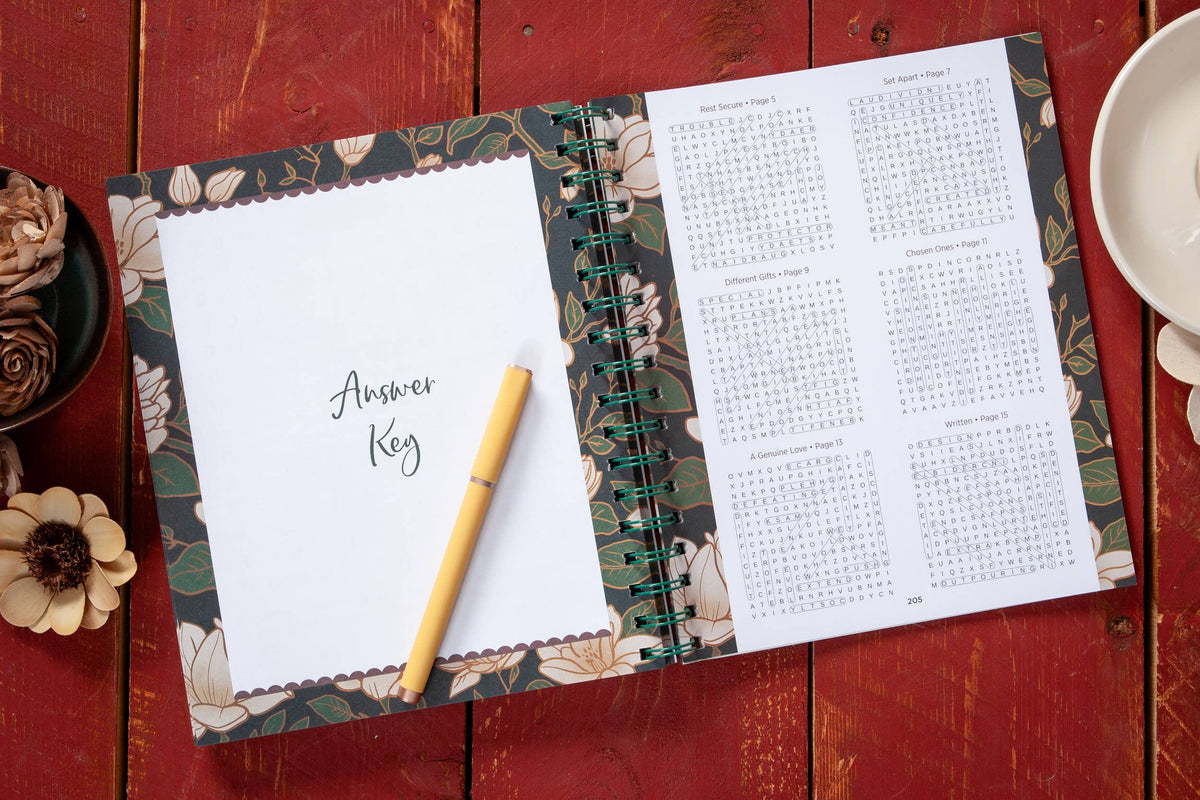 An open spiral-bound copy of the “100 Days of Strength for Women Word Search Book” by BroadStreet Publishing Group, LLC rests on a red wooden table, displaying an Answer Key page with a yellow pen and floral design beside solution pages featuring answer grids. Nearby sit a white cup and artificial flowers.
