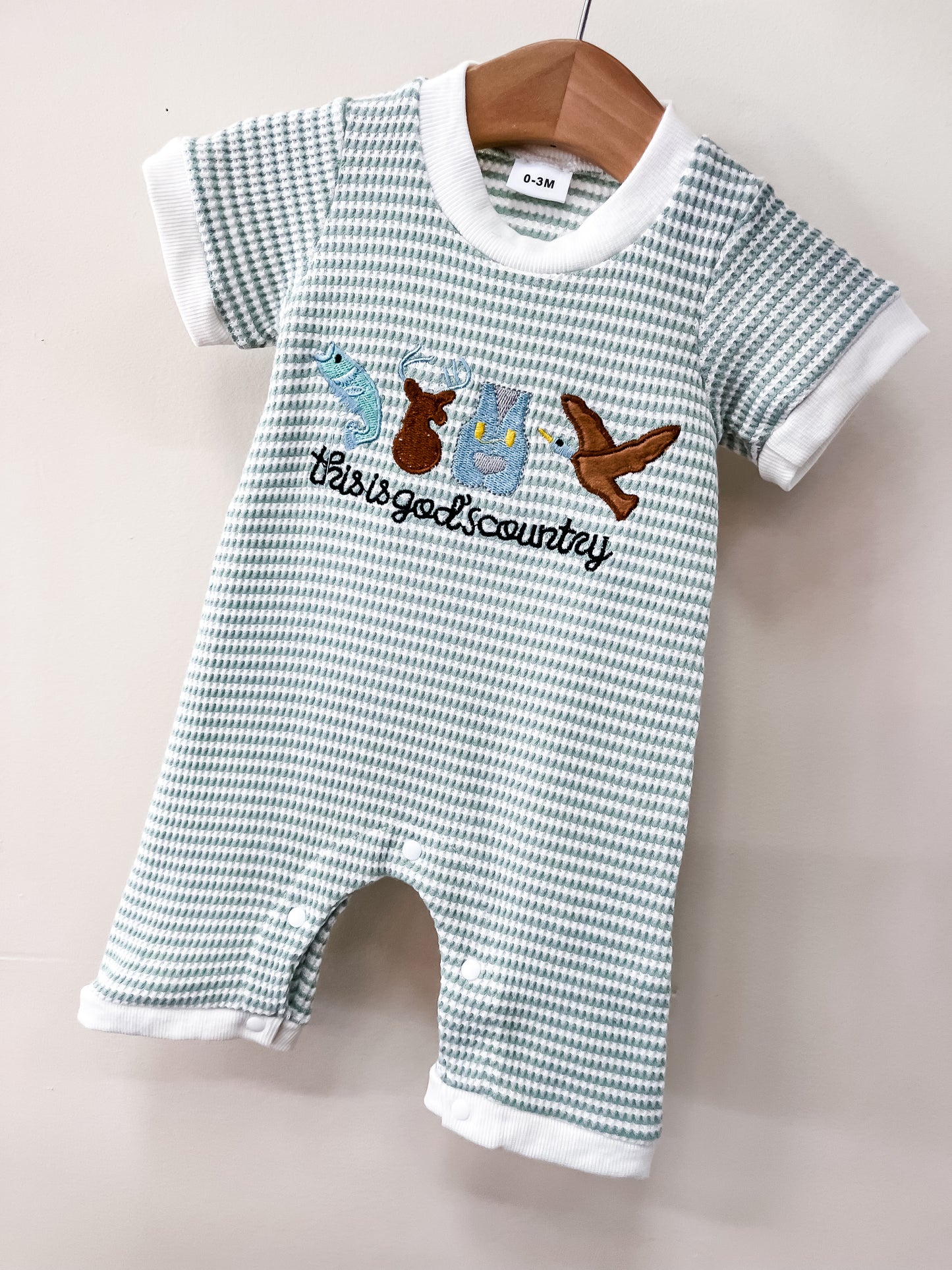 The God's Country Romper by Tiny Tot Bundles features a green and white checkered pattern, embroidered animals (snake, kangaroo, koala, bird), and "this is gods country" text on the front, displayed on a wooden hanger.