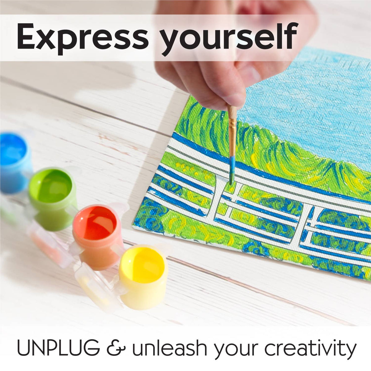 A hand brings The Japanese Footbridge to life with blue and green hues on an artist canvas from the Paint by Number Museum Series. Four small Faber-Castell paint pots sit nearby. Text reads: Express yourself. UNPLUG & unleash your creativity—paint like the Masters with Creativity for Kids.