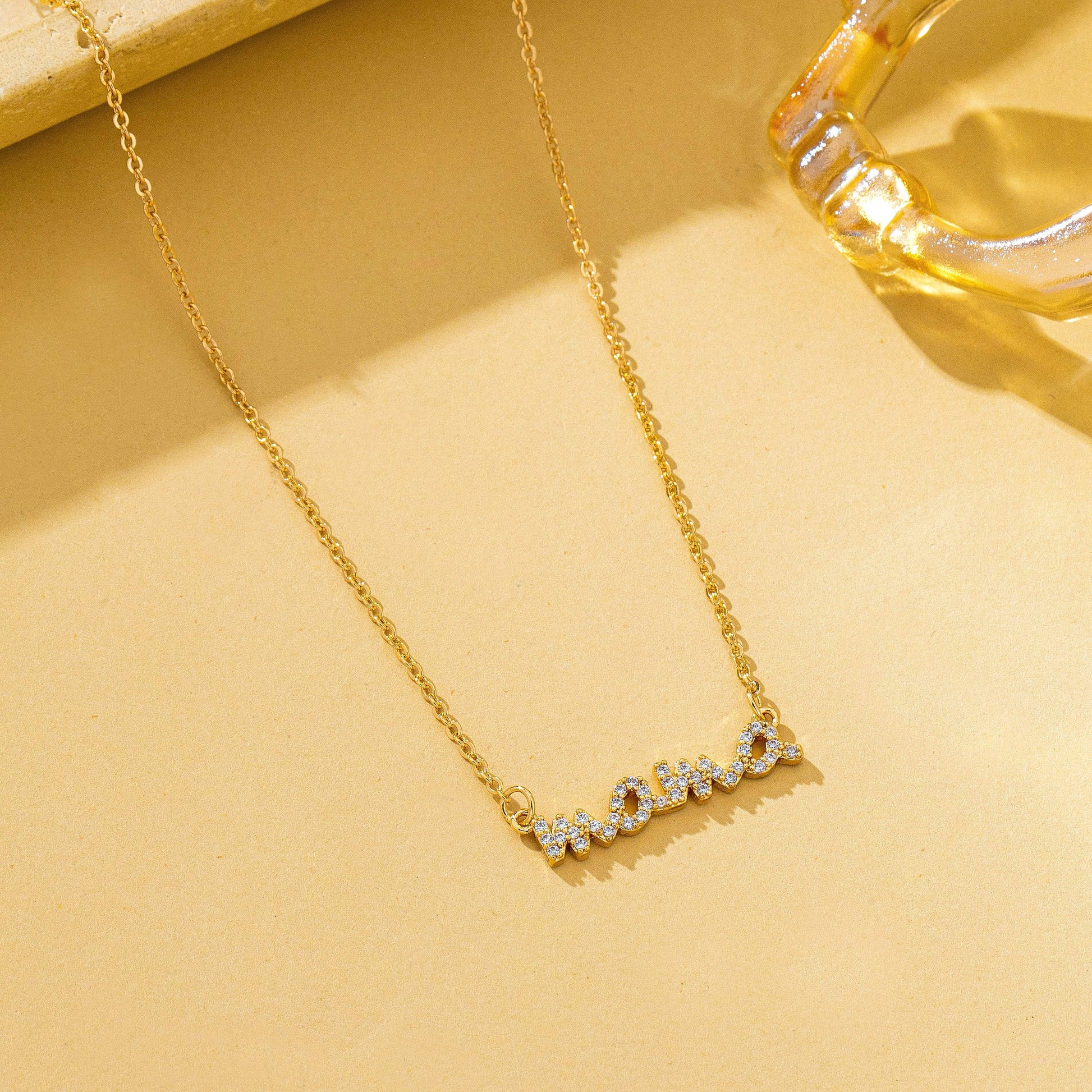 The JOSSLYN by wall to wall Gold MAMA Necklace, featuring a cursive pendant adorned with small sparkling stones, is displayed on a beige surface beside a transparent, light gold glass object.