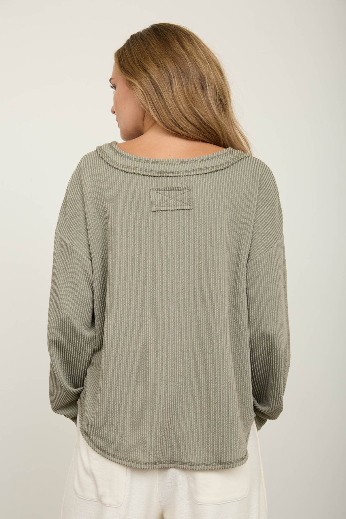A woman with long, light brown hair is shown from the back wearing the Blu Pepper Olive V-Neck Long Sleeve Top and white pants. The background is plain and light-colored.