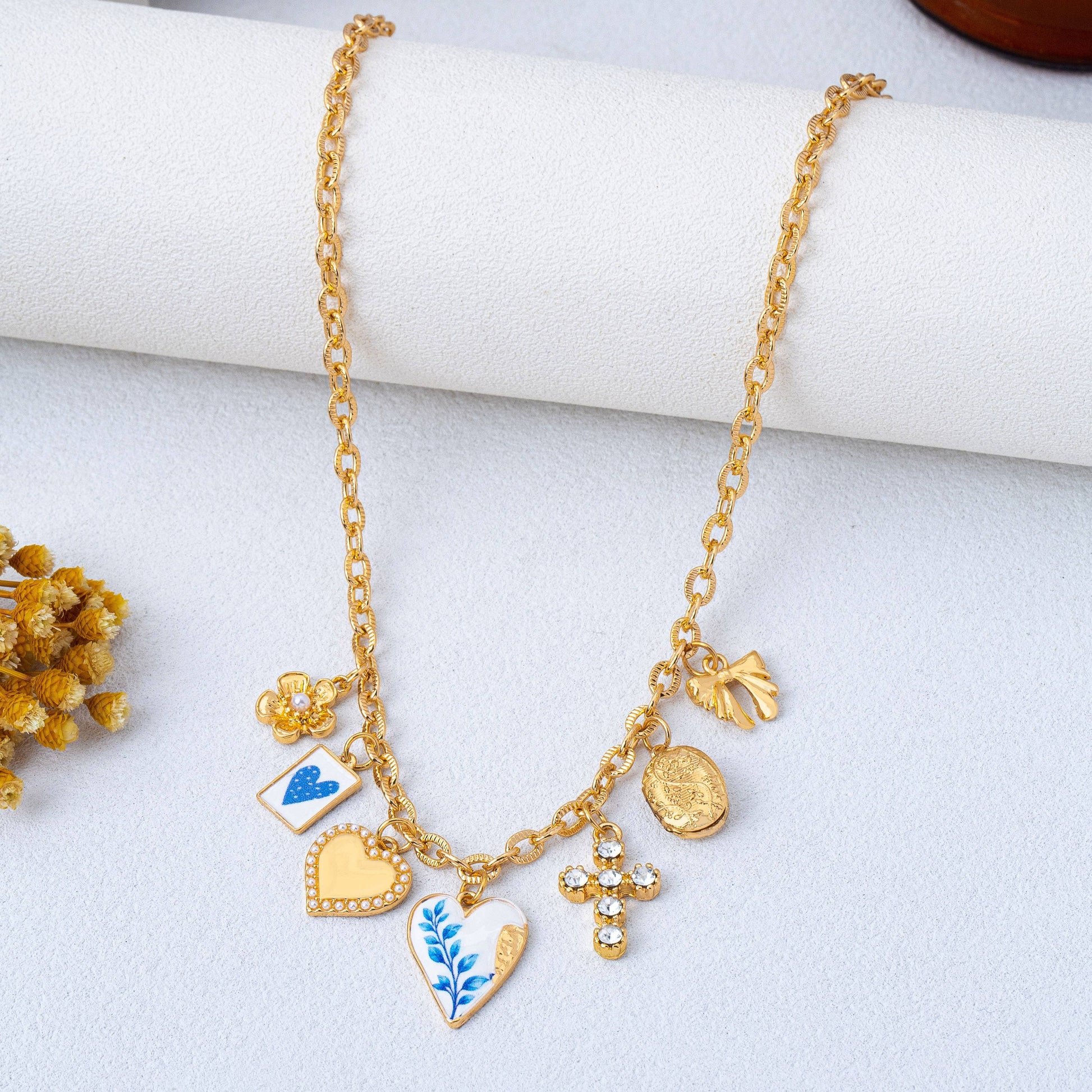 The Blue Vintage Coquette Charm Necklace by JOSSLYN by wall to wall features a gold chain adorned with charms—including a blue patterned heart, gold heart, flower, gem-studded cross, medallion, and small gold hand—elegantly displayed on a white surface with dried flowers nearby.