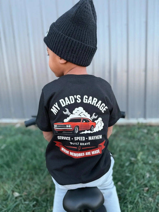 A child wearing the Raising Brave My Dad's Garage Toddler Tee—a black shirt featuring a red car graphic and the text “MY DAD’S GARAGE—Service, Speed, Mayhem—Where memories are made”—and a black beanie sits on a bike facing away from the camera.