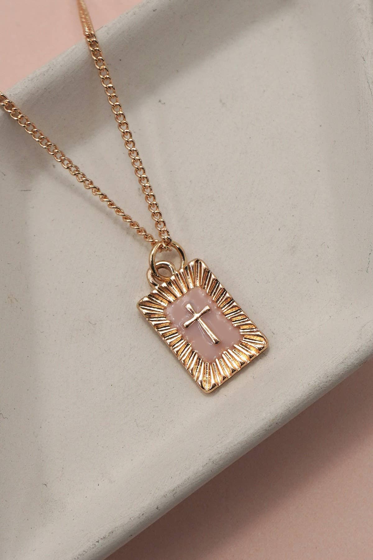 The Sunburst Square Cross Pendant Necklace by JOSSLYN by wall to wall features a rectangular gold pendant with a pink center and a small gold cross, displayed on a white ceramic tray against a light pink background.