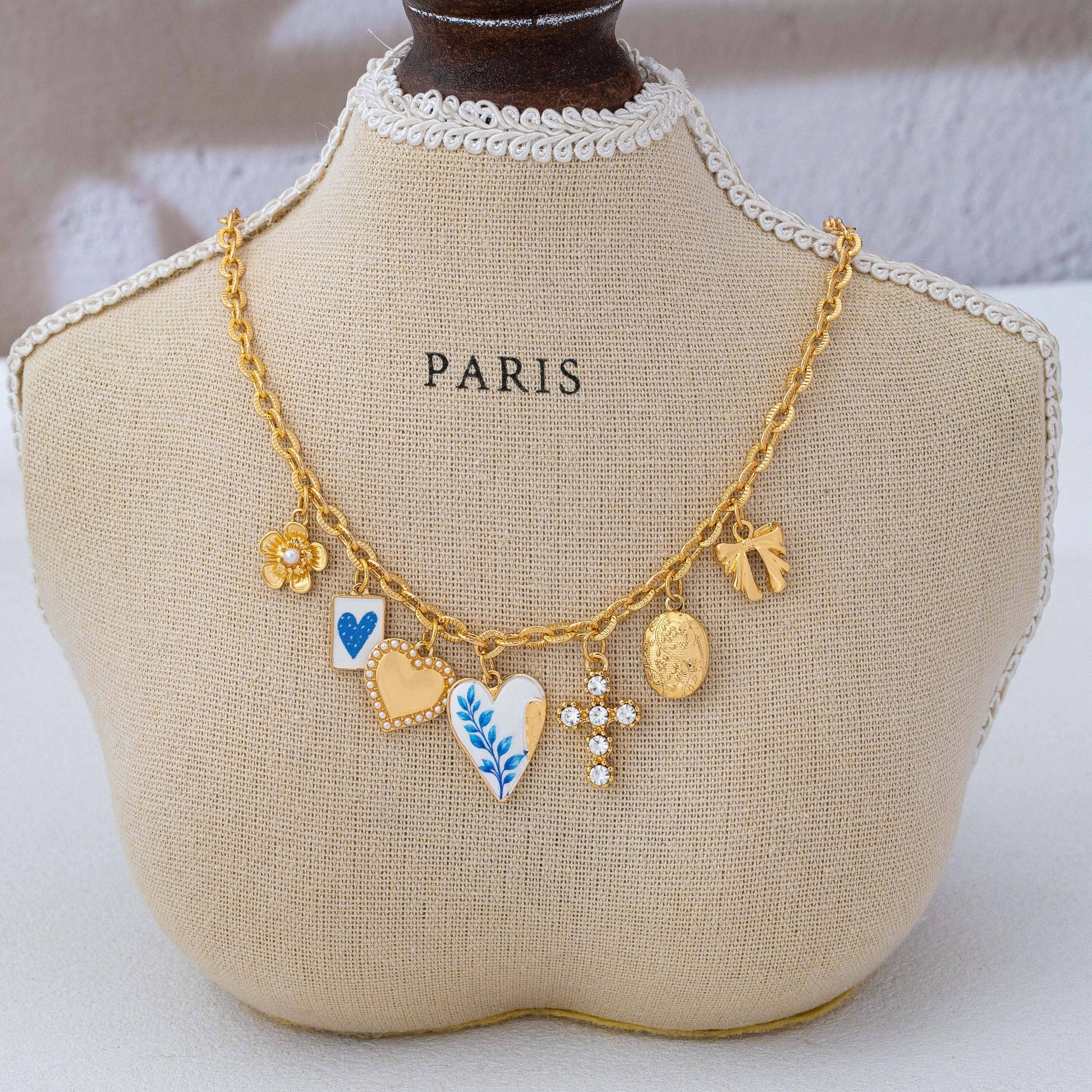 A beige mannequin bust with “PARIS” printed on it displays the Blue Vintage Coquette Charm Necklace by JOSSLYN by wall to wall—a gold chain adorned with seven charms, including a flower, heart, leaf, blue-white enamel, medallion, and daisy shapes.