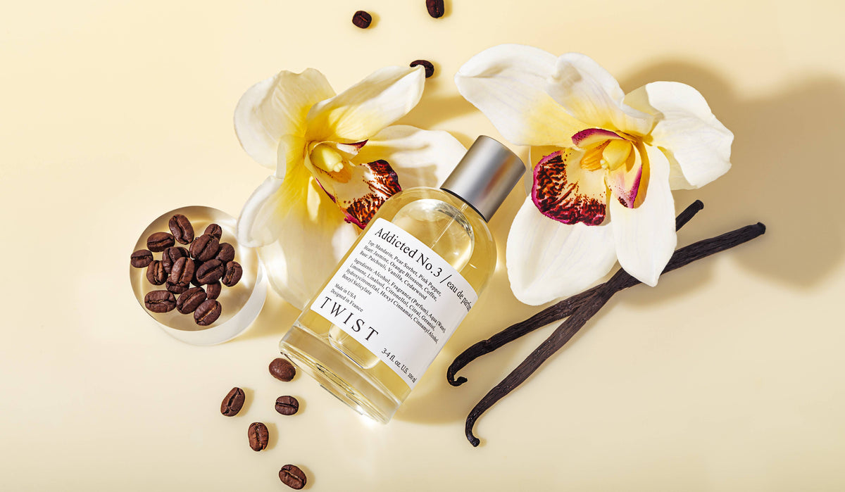 A bottle of Addicted No.3 Perfume by House of Twist rests amid two white orchids, coffee beans, vanilla pods, and a small silver dish of coffee beans on a light beige surface—embodying the essence of a French fragrance in this long-lasting women’s perfume.