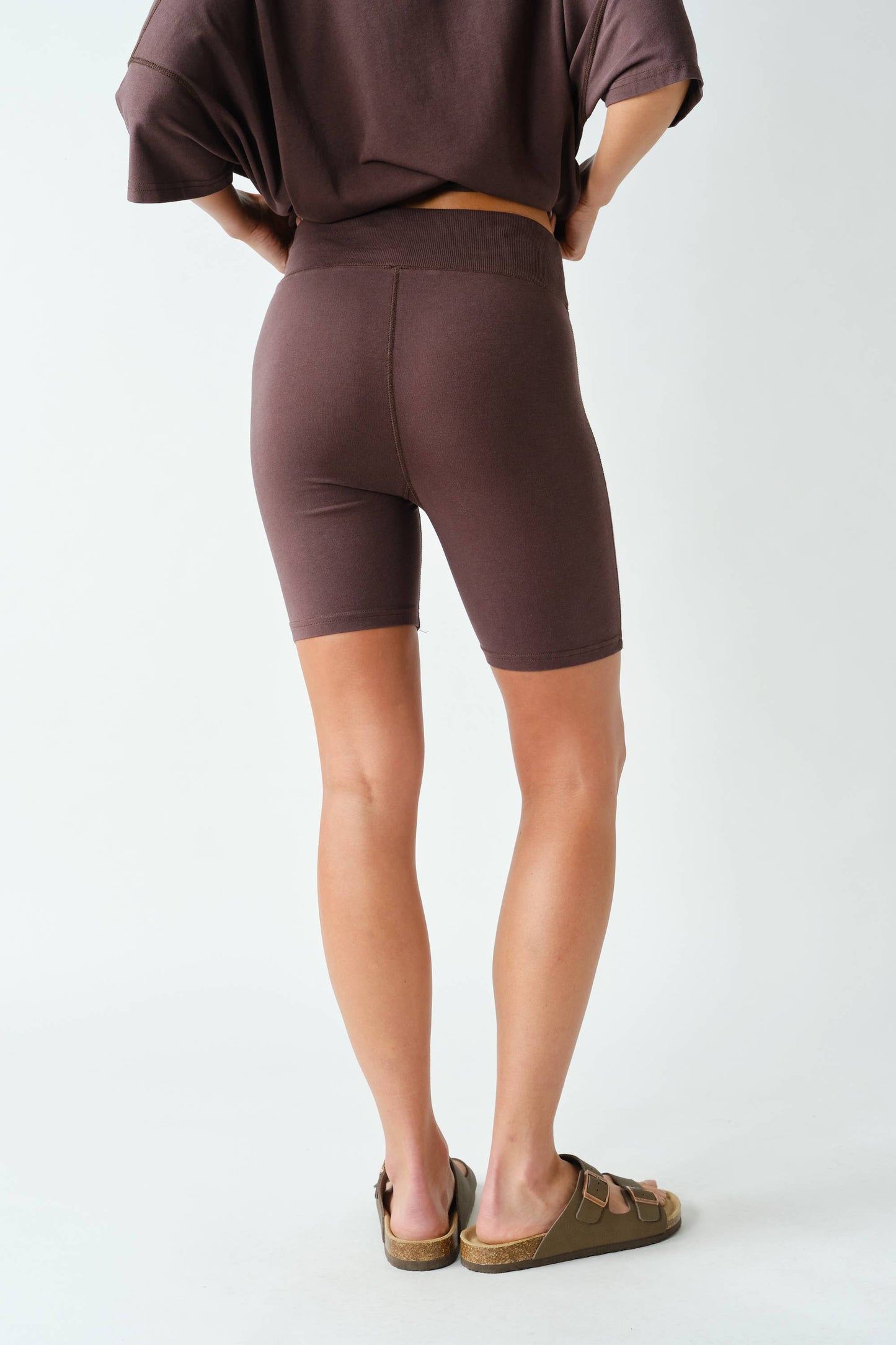 A person stands with their back to the camera, wearing the Mono B Switchback Pima Cotton Set—featuring a brown short-sleeve top and matching fitted shorts—with tan sandals, set against a plain white background.