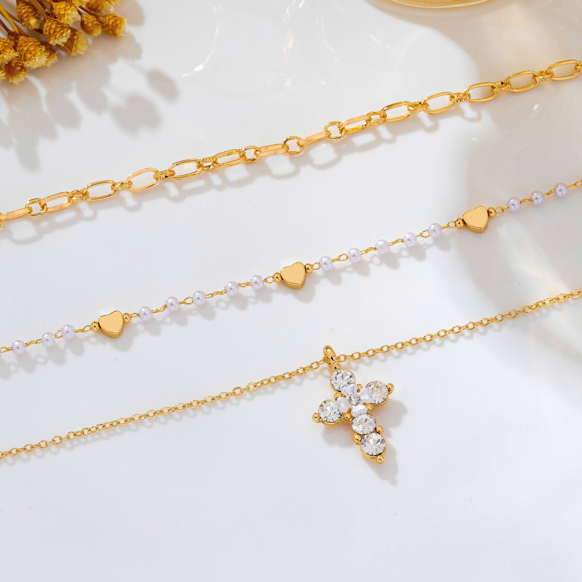 Discover the JOSSLYN by wall to wall Multi-Strand Pearl & Crystal Cross Necklace, featuring three delicate gold chains—one with elongated links, one adorned with small heart and bead accents, and one showcasing a sparkling crystal cross pendant—all elegantly displayed on a white surface beside dried flowers.