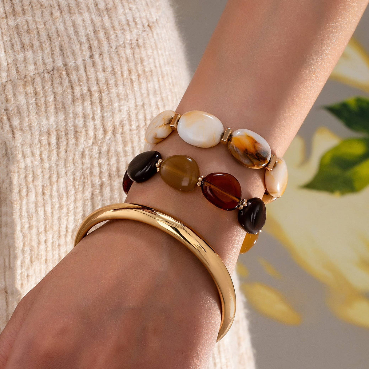 A close-up of a person’s wrist adorned with the JOSSLYN by wall to wall Boho Gold Bracelet Set—featuring two earth-toned beaded gemstone bracelets and a shiny gold bangle—set against a softly blurred floral background. The individual wears a textured beige sweater.