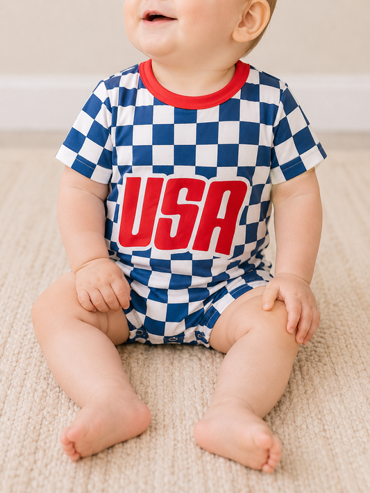 A baby sits on the floor, smiling and partially out of frame, wearing the Tiny Tot Bundles USA Check Bubble— a blue and white checkered romper with red trim and bold red "USA" letters on the front.