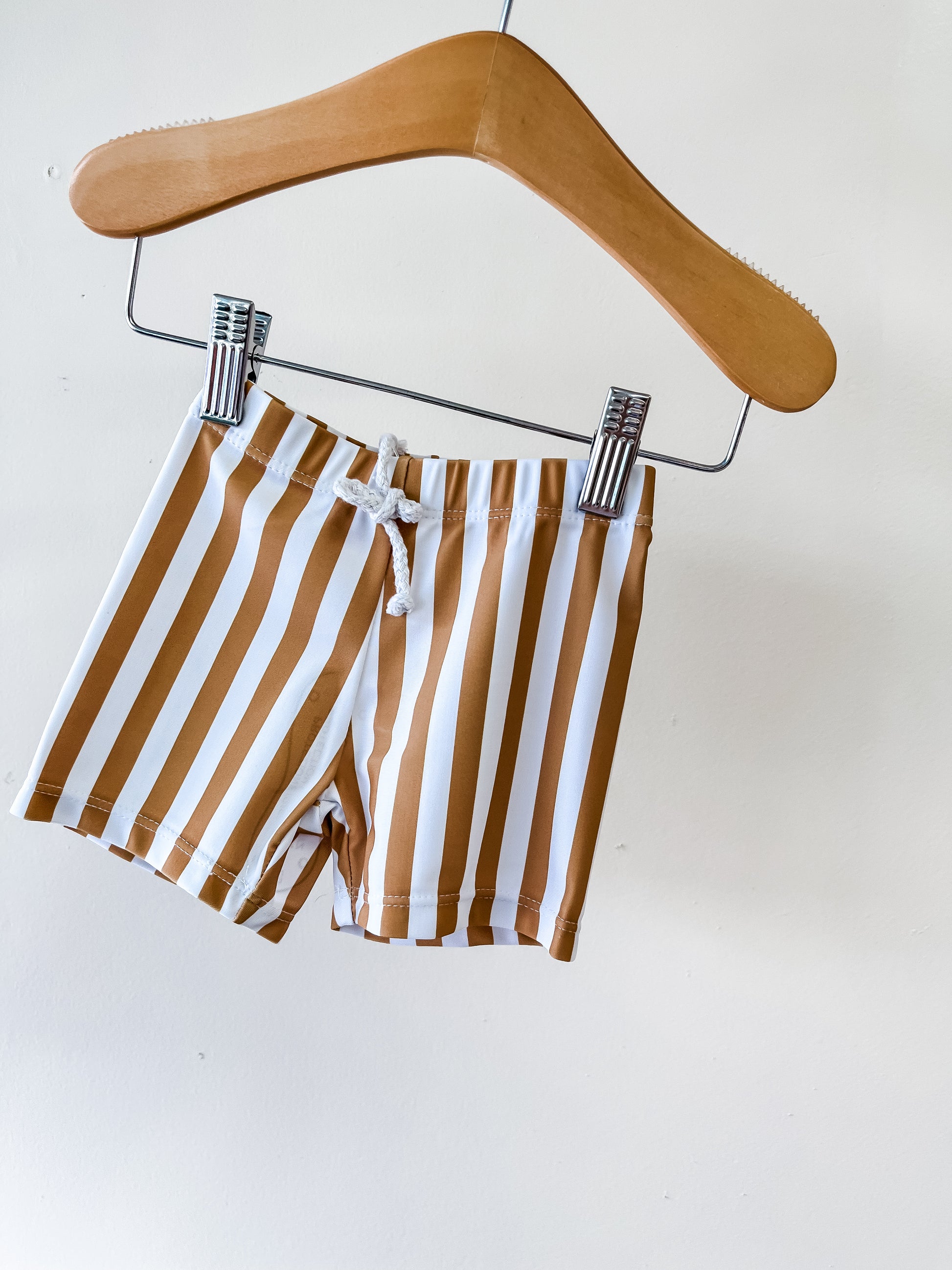 The Caramel Stripe Swim Shorts by Tiny Tot Bundles, featuring vertical brown and white stripes, hang on a wooden hanger against a plain white background.