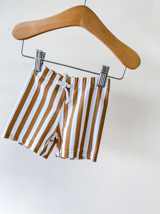 The Caramel Stripe Swim Shorts by Tiny Tot Bundles, featuring vertical brown and white stripes, hang on a wooden hanger against a plain white background.