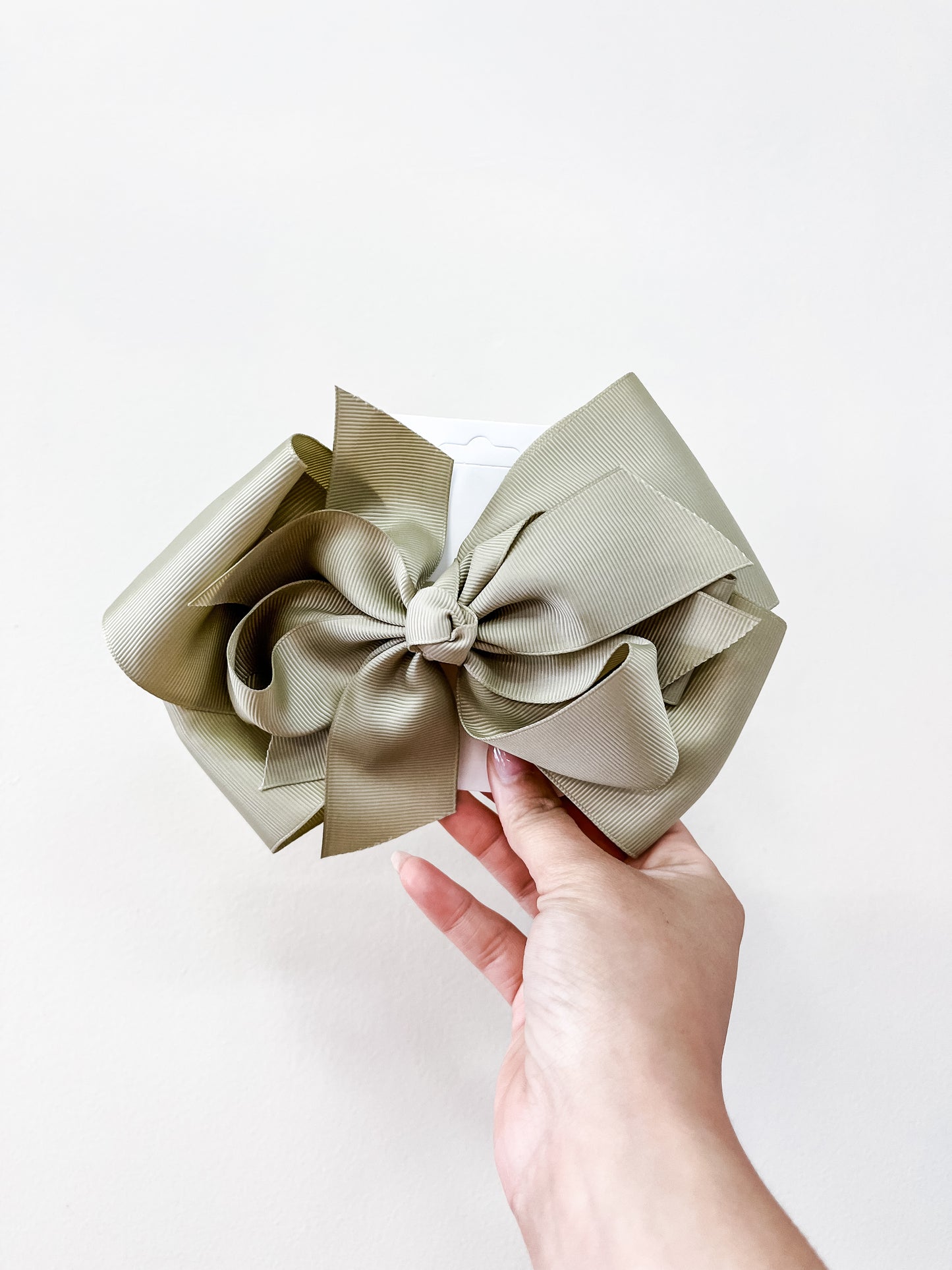 A hand holds a large pastel olive green Oversized Pastel Bow by funteze, made from textured fabric, against a plain white background.