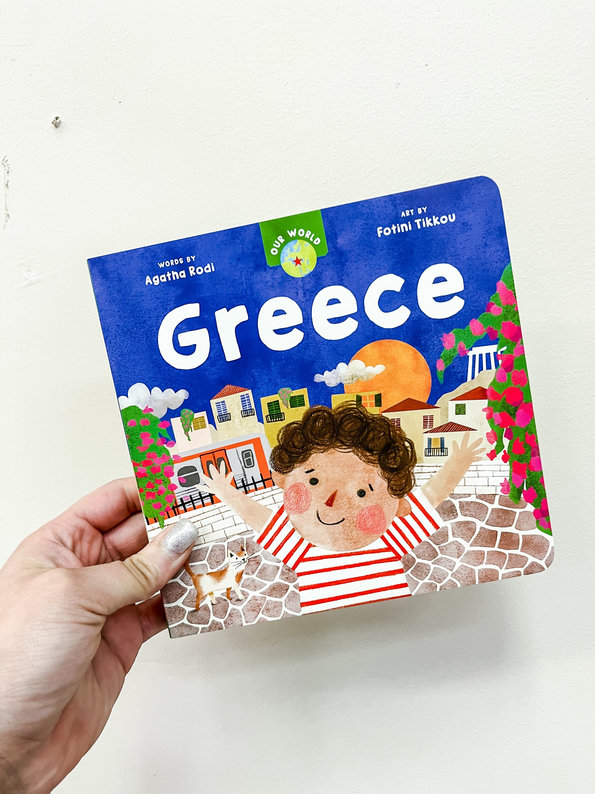 A hand holds Barefoot Books' "Our World: Greece Book," a colorful children’s book featuring a smiling child, white houses, pink flowers, an orange sun, with words by Agatha Rodi and art by Fotini Tikkou.