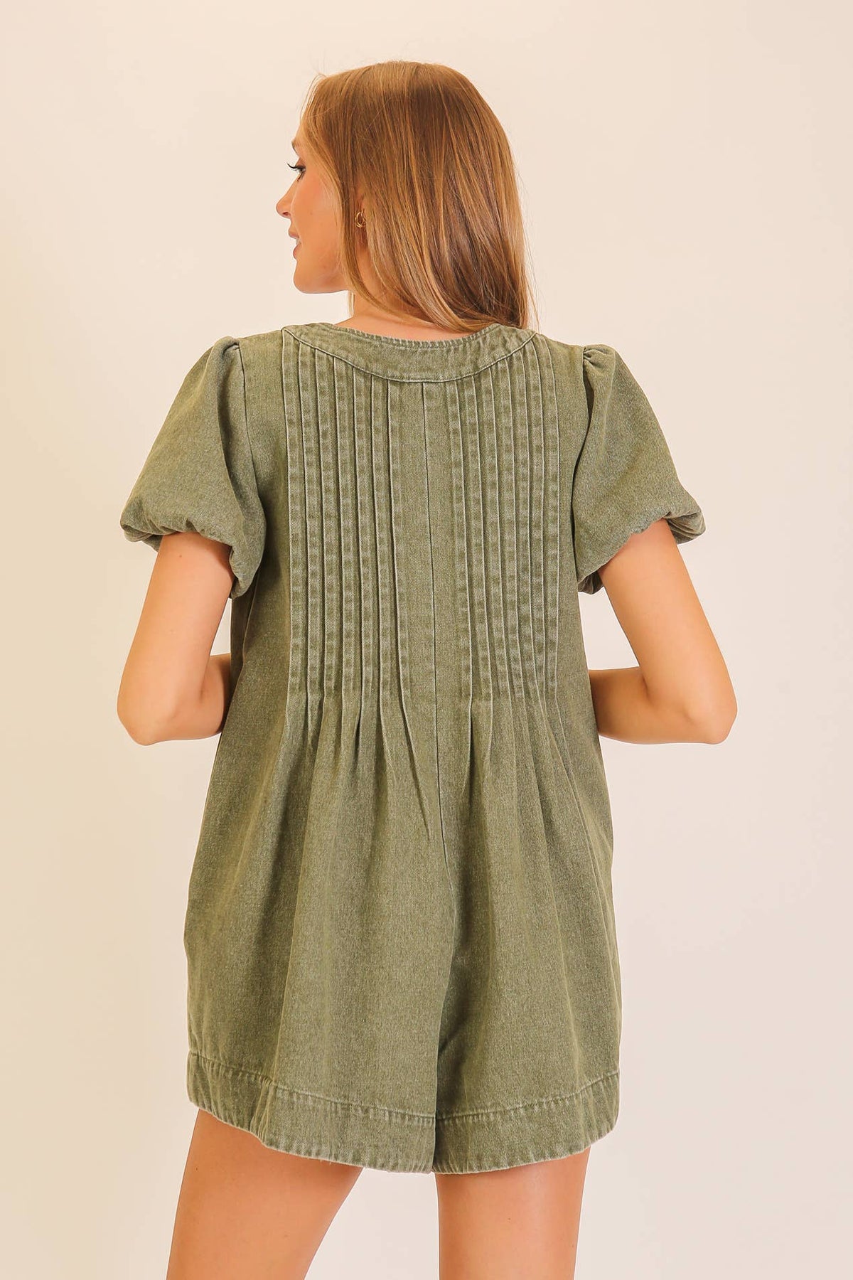 A woman with long light brown hair wears the Lumiere Desert Muse Utility Romper, featuring short sleeves and pleated back details, as she stands against a plain light background.