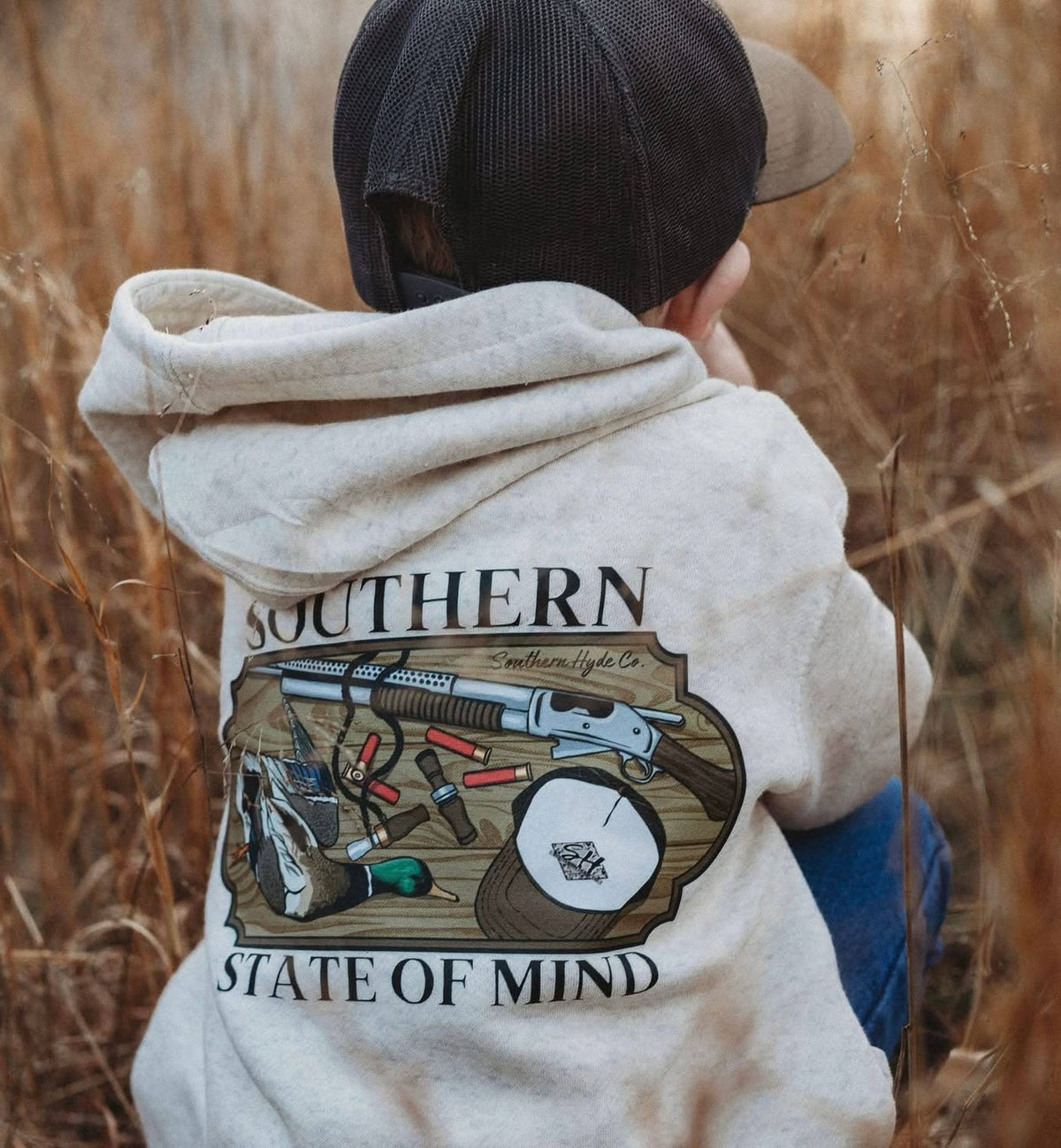 Child wearing a beige Southern State of Mind Hoodie featuring a graphic of a shotgun, shotgun shells, a duck, and a baseball cap on the back.