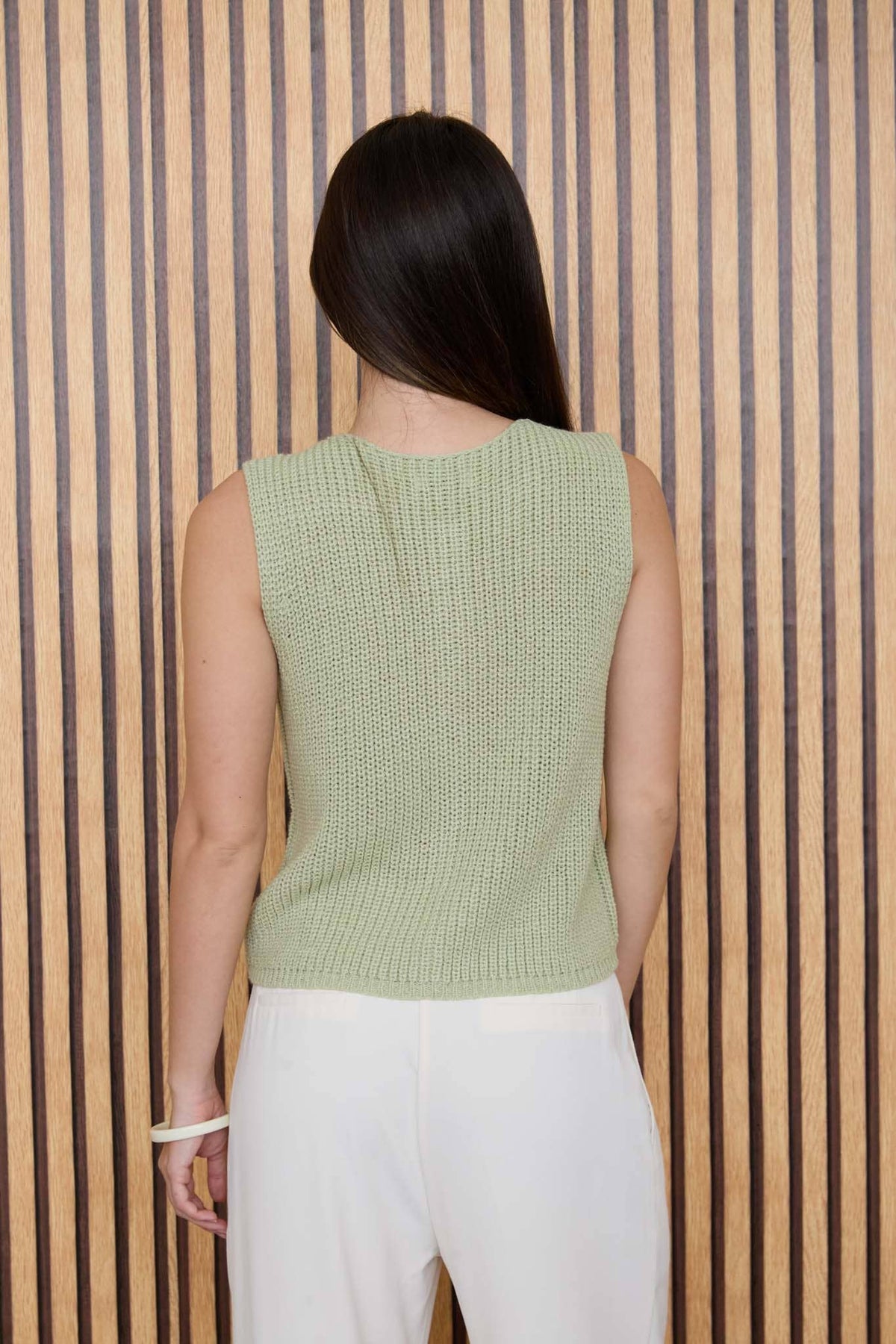 A person with long dark hair, wearing The Workshop Studio’s Sage Knit Button Down Cardigan and white pants, stands facing a vertical striped wooden wall.