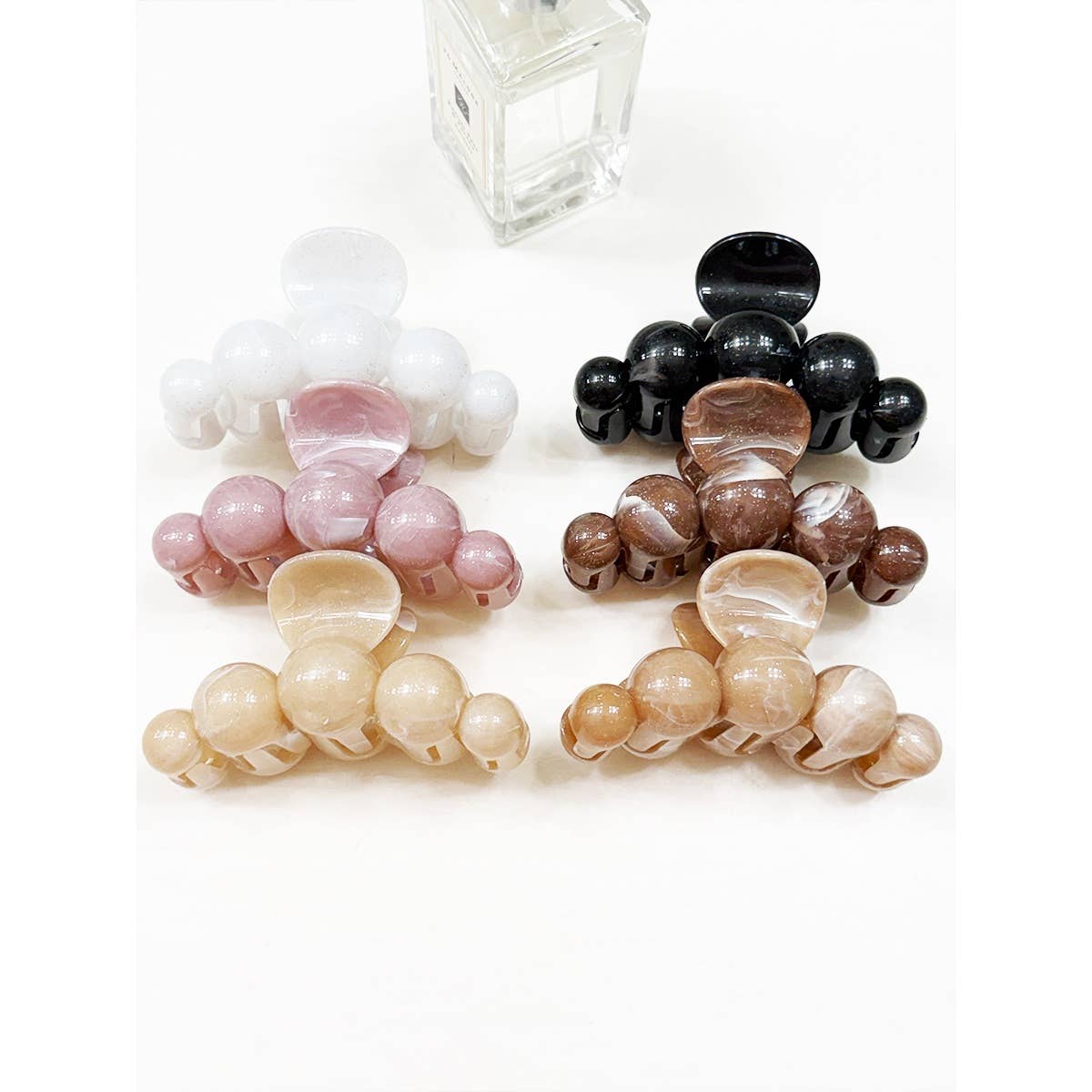 Four Love and Repeat Large Marbled Hair Claws are arranged on a white surface in shades of white, black, pink, brown, and beige. A small glass perfume bottle sits in the background.
