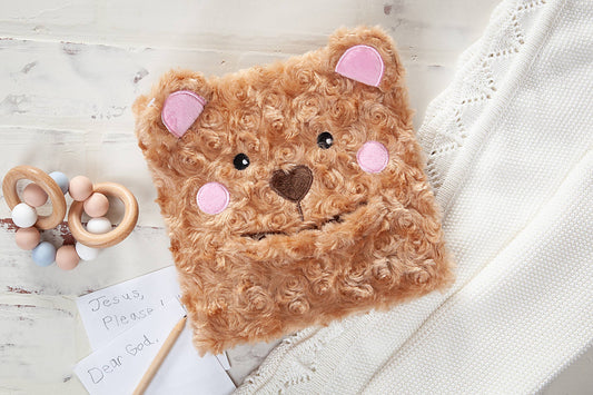 A Prayer Bear Book by BroadStreet Publishing Group, LLC rests on a white knit blanket next to wooden baby toys and a handwritten note reading “Jesus, please” and “Dear God.”.
