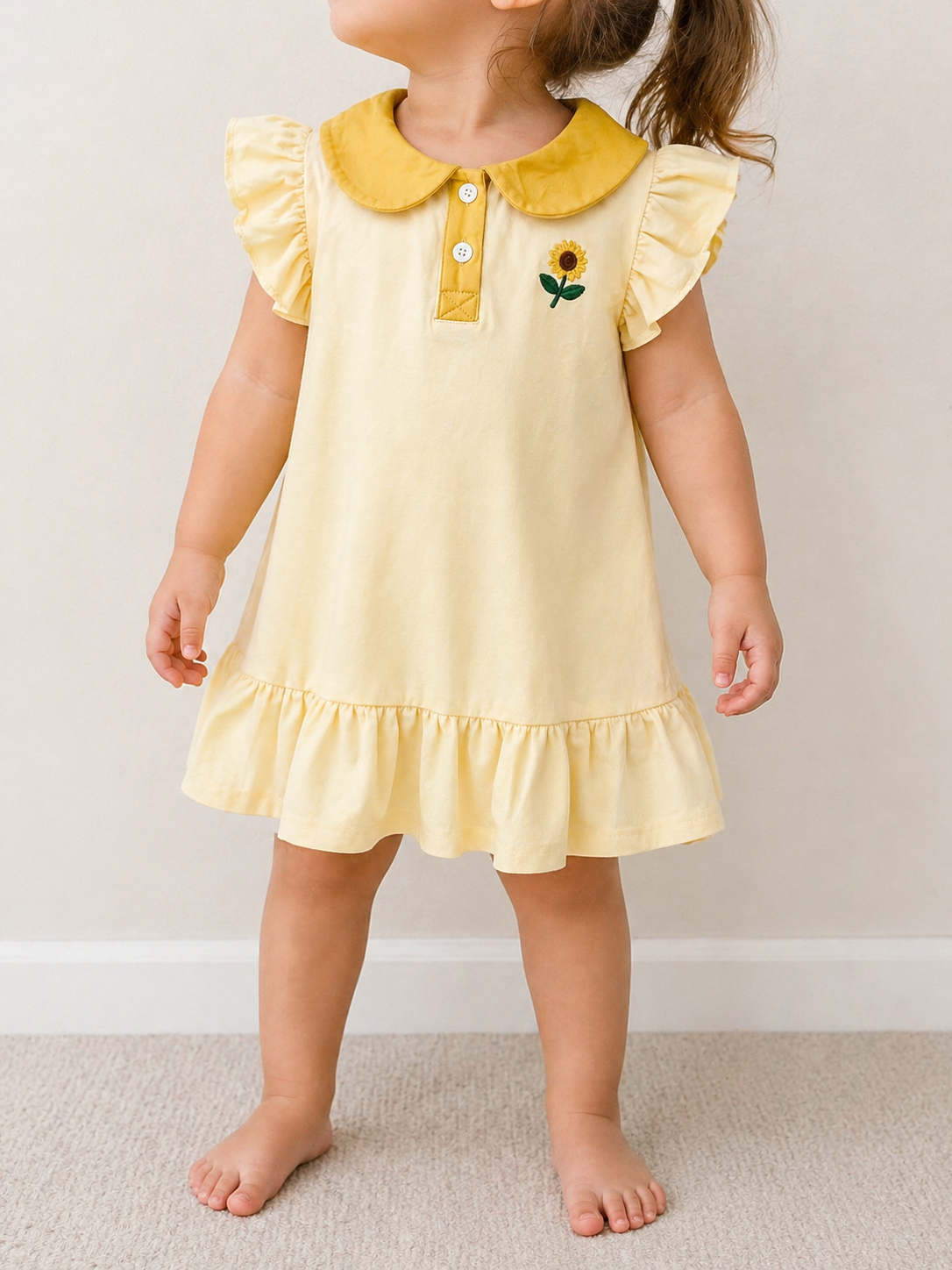 A young child is barefoot indoors, wearing the Sunflower Ruffles Dress by Tiny Tot Bundles—a light yellow dress with ruffled sleeves and hem, a yellow collar with white buttons, and a small sunflower embroidered on the chest.
