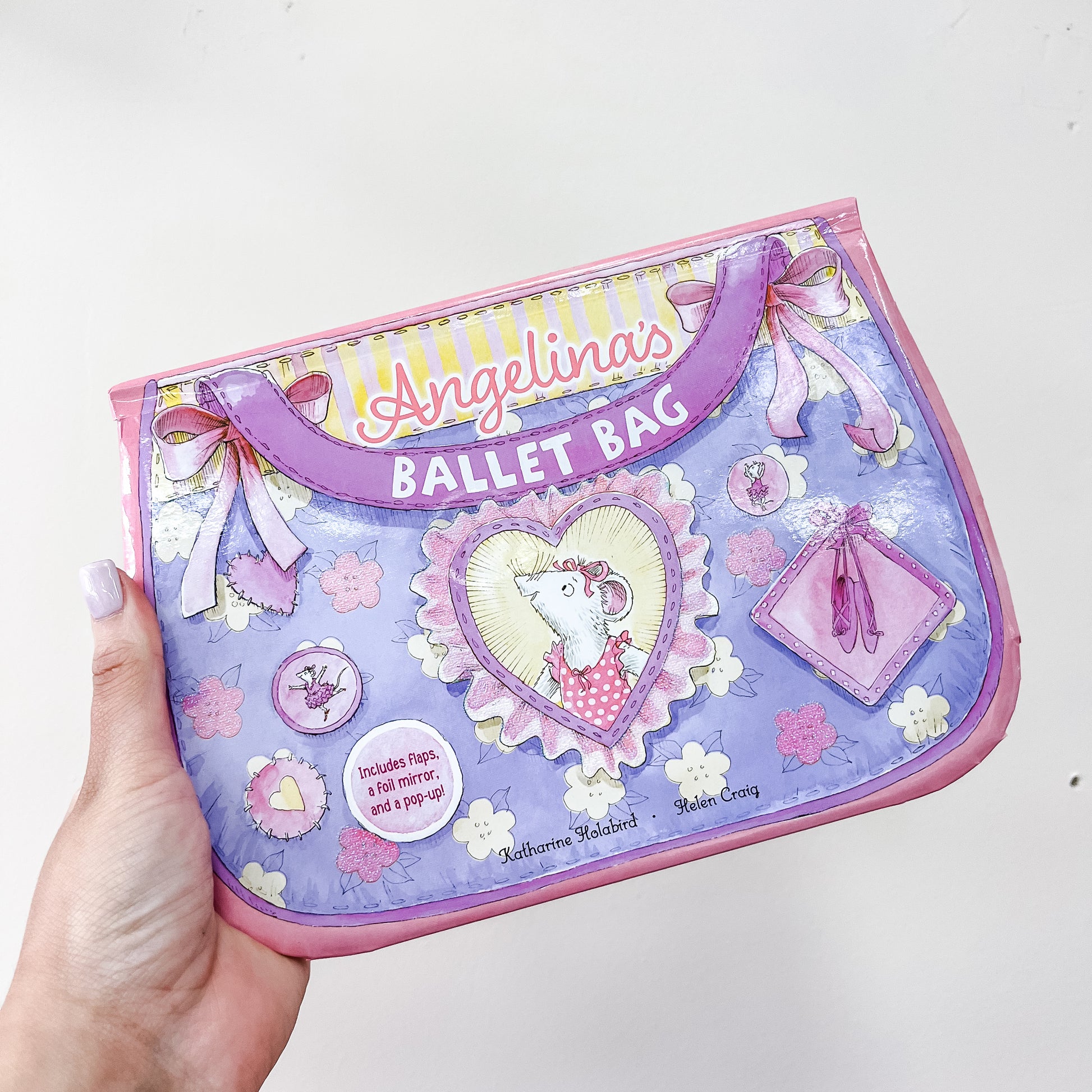 A hand holds Simon & Schuster's "Angelina's Ballet Bag Book," a purple and pink children's book featuring bows, ballet slippers, flowers, and a mouse in a tutu inside a heart-shaped frame.