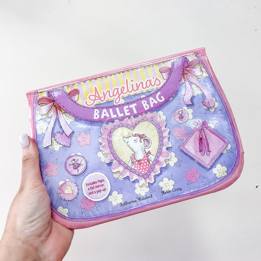 A hand holds Simon & Schuster's "Angelina's Ballet Bag Book," a purple and pink children's book featuring bows, ballet slippers, flowers, and a mouse in a tutu inside a heart-shaped frame.