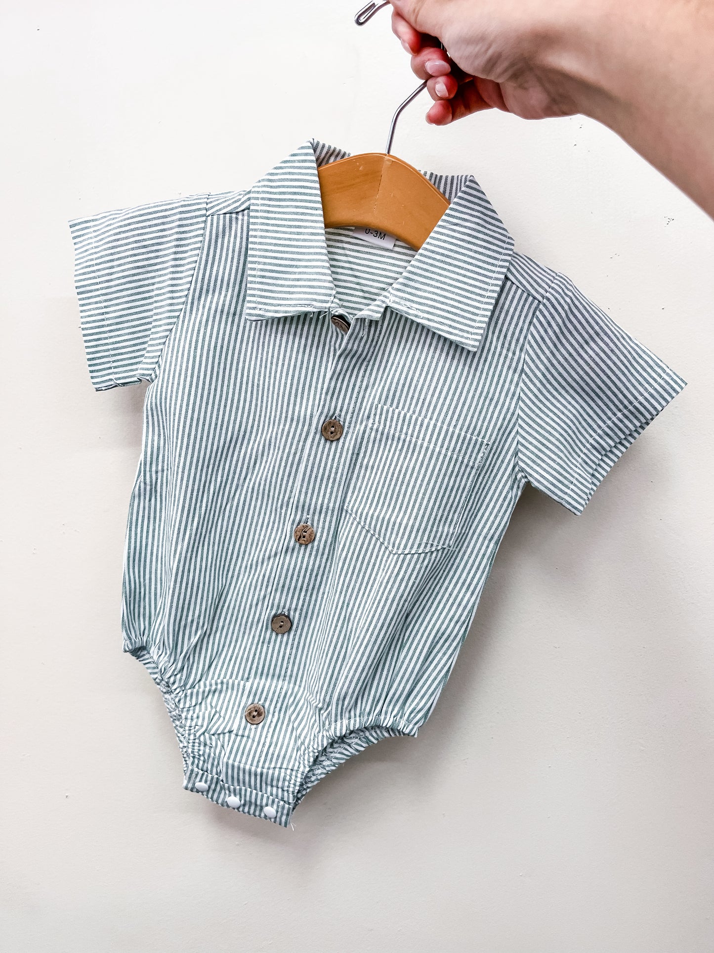 A hand holds Tiny Tot Bundles' Green Stripe Collared Bubble— a baby romper with green and white stripes, brown buttons, a collar, and a front pocket—pictured on a hanger against a plain white background.