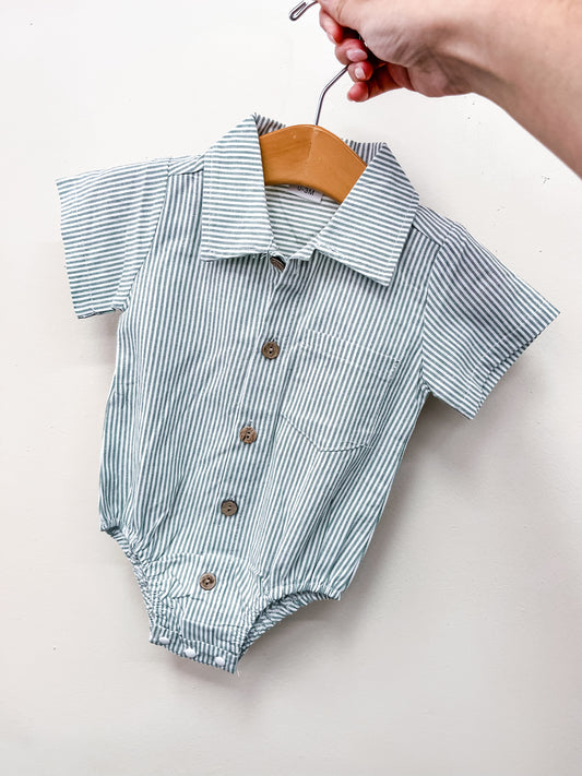 A hand holds Tiny Tot Bundles' Green Stripe Collared Bubble— a baby romper with green and white stripes, brown buttons, a collar, and a front pocket—pictured on a hanger against a plain white background.