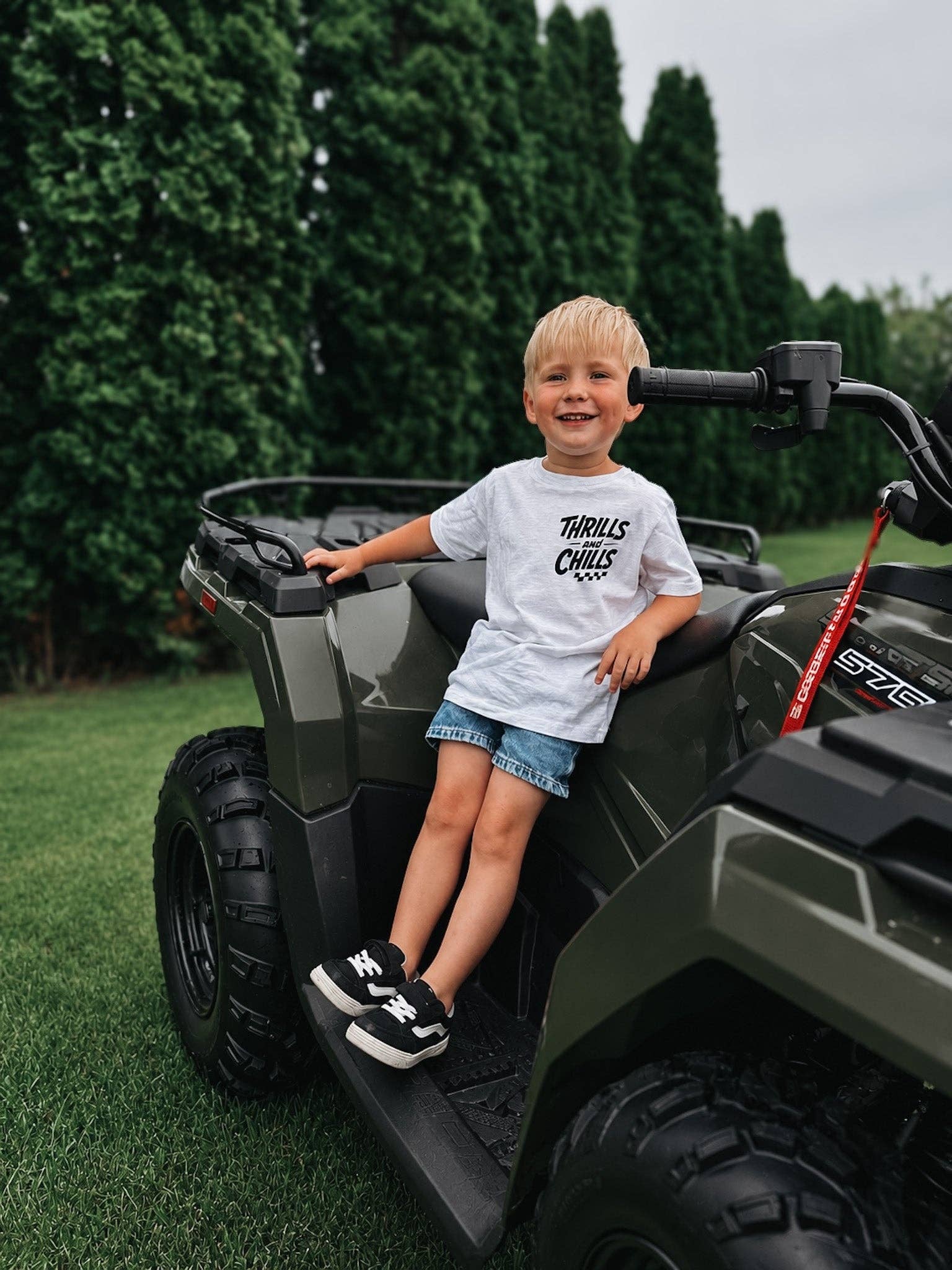 A young boy in a Raising Brave Big Thrills and Chills Toddler Tee sits smiling on a dark green ATV, with tall green trees and grass in the background.