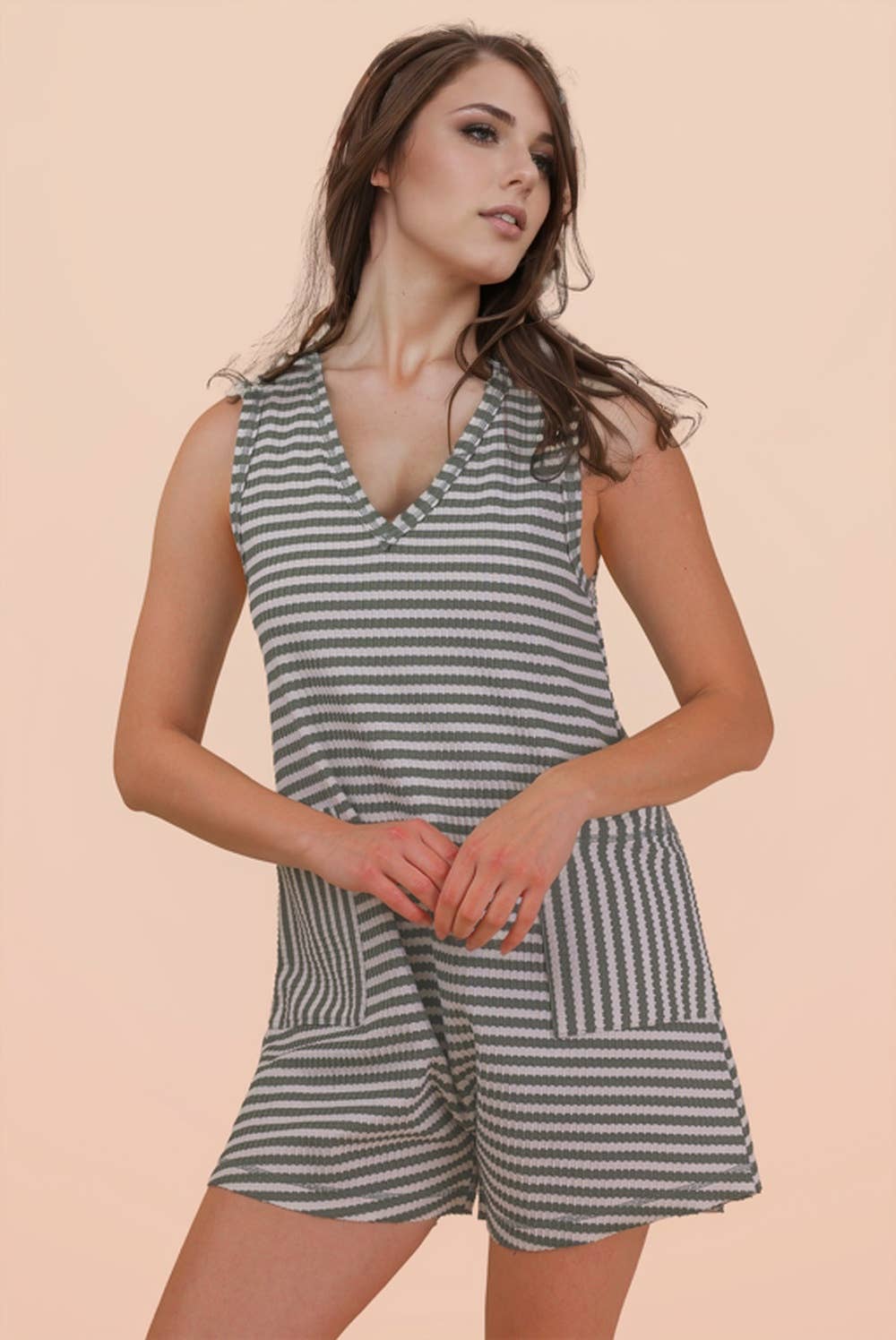 A woman models the VERY J Striped Knit Casual Romper, featuring a sleeveless design, gray and white stripes, and front pockets, against a plain peach background.
