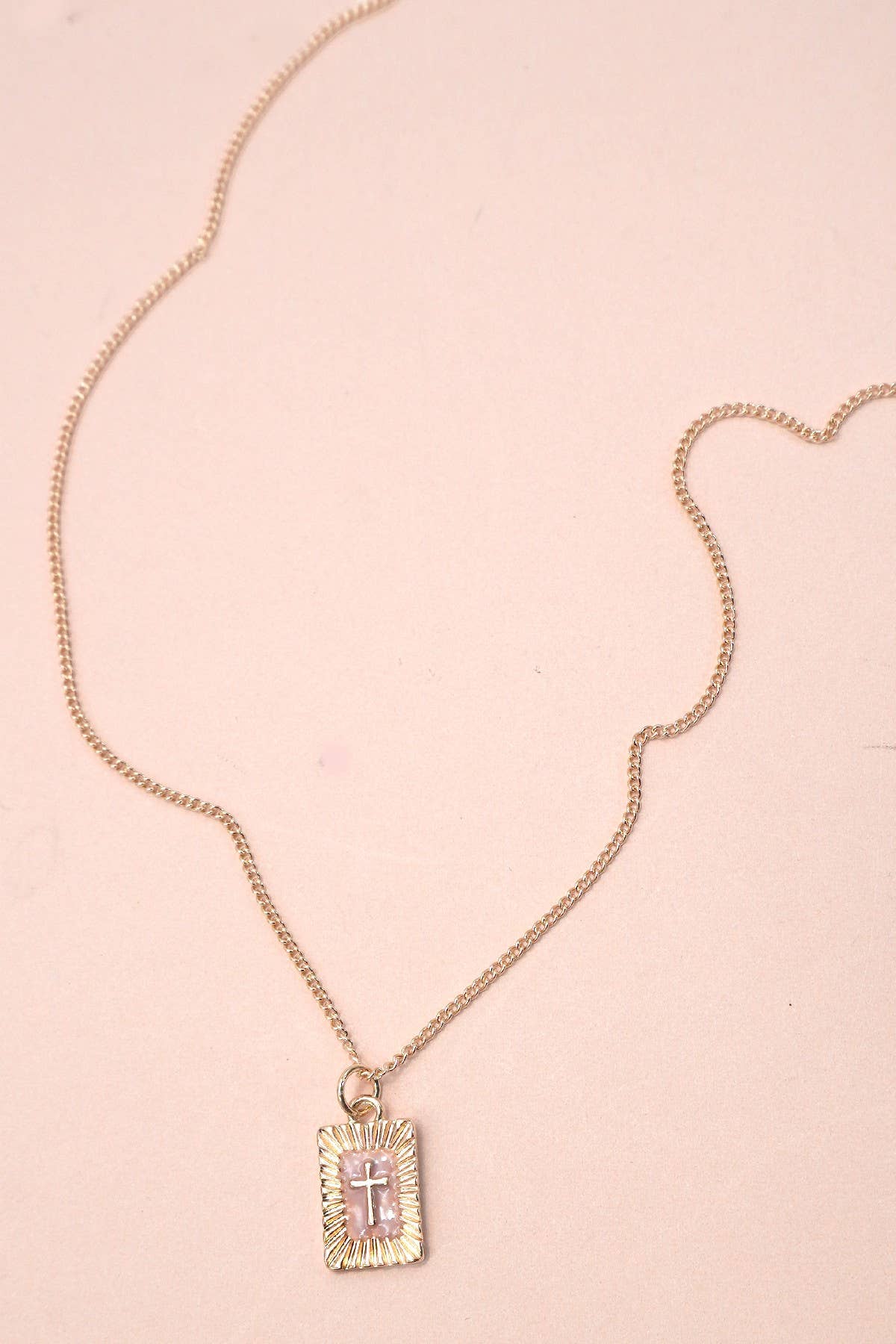 JOSSLYN by wall to wall’s Sunburst Square Cross Pendant Necklace is shown on a pale pink background, featuring a gold chain and a rectangular pendant with a raised cross design.