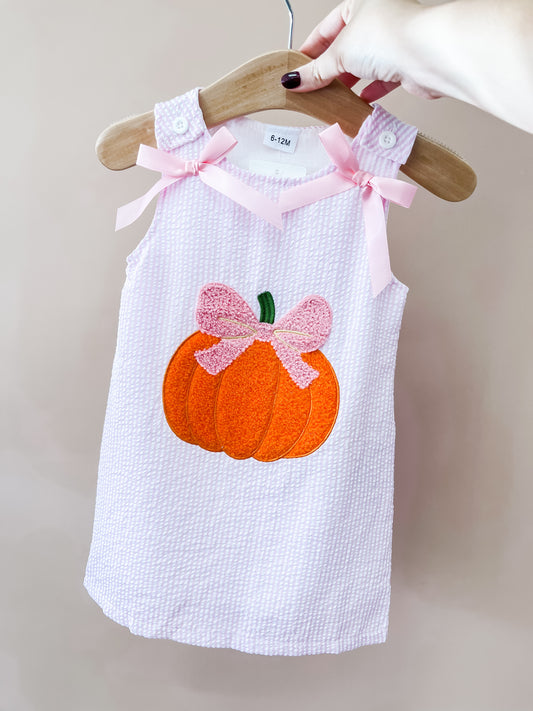 The Tiny Tot Bundles Pink Bow Pumpkin Dress is a pink toddler dress featuring pink bows on the shoulders and an orange pumpkin with a pink bow applique on the front, displayed on a wooden hanger.