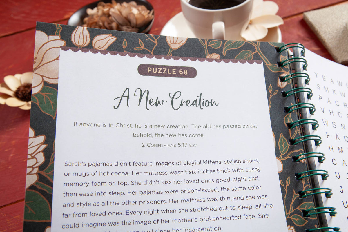 A close-up of the 100 Days of Strength for Women Word Search Book by BroadStreet Publishing Group, LLC open to “A New Creation,” featuring a verse from 2 Corinthians 5:17. A teacup, flower, and pen sit nearby on a red table.