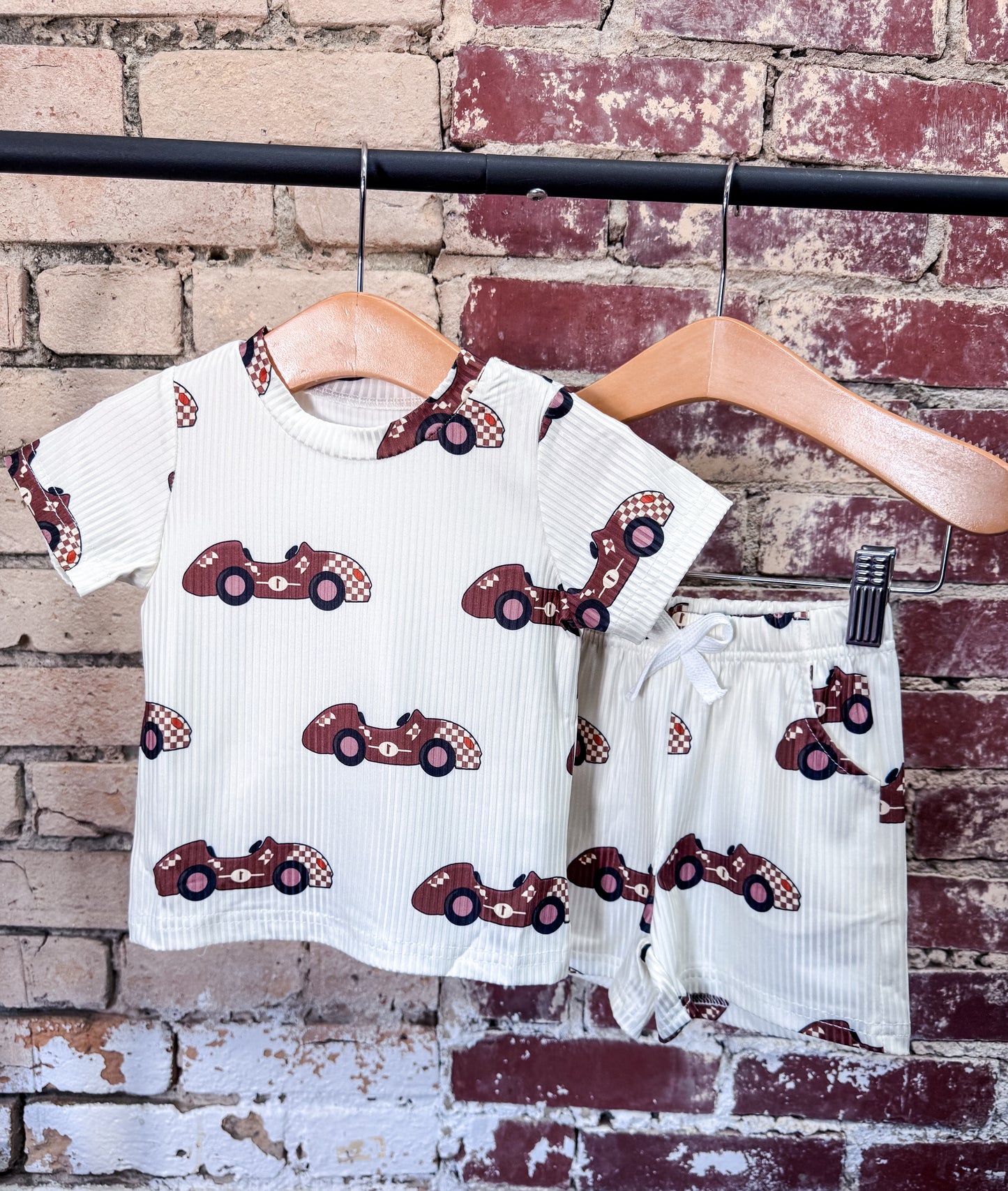 The Tiny Tot Bundles Racecar Driver Boy Set is a children’s matching T-shirt and shorts outfit with a red racecar print, displayed on wooden hangers against a rustic brick wall.