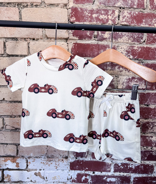 The Tiny Tot Bundles Racecar Driver Boy Set is a children’s matching T-shirt and shorts outfit with a red racecar print, displayed on wooden hangers against a rustic brick wall.