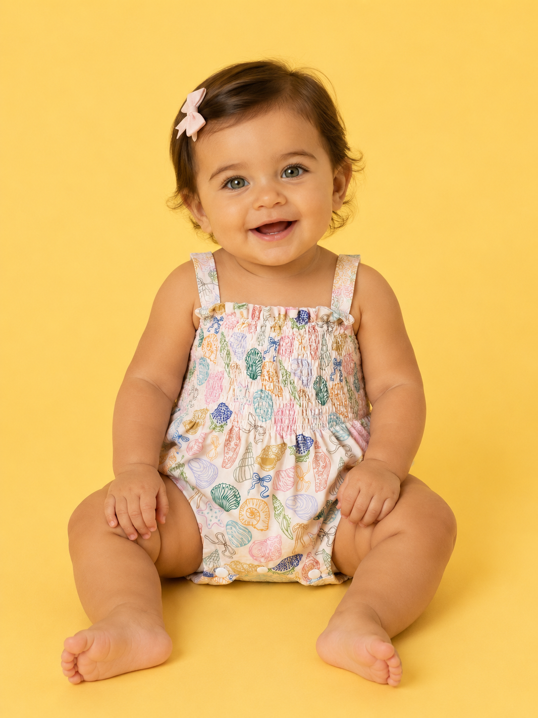 A smiling baby with light brown hair and a pink bow sits on a yellow background, wearing the Treasure Island Smocked Romper by Tiny Tot Bundles.