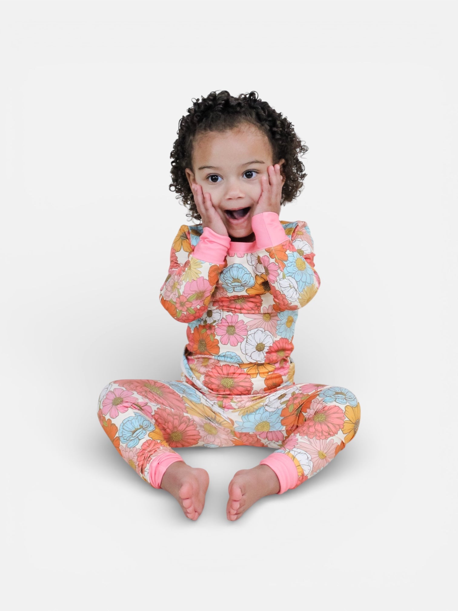 A young child with curly hair sits on the floor on a plain white background, looking surprised and happy while wearing the colorful Retro Bloom Jammies Set by Tiny Tot Bundles.