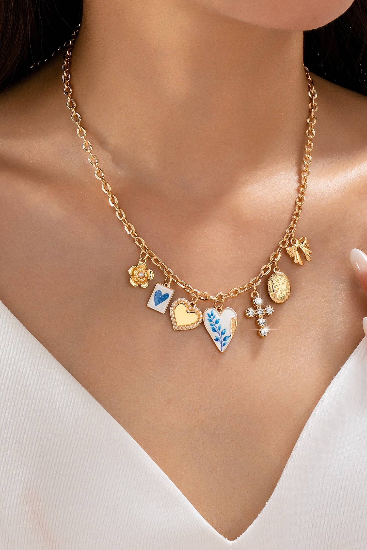 Wearing a white v-neck top, the person showcases the JOSSLYN by wall to wall Blue Vintage Coquette Charm Necklace, a gold chain adorned with six unique charms—hearts, a flower, a blue leaf design, a coin, a bow, and a cross.