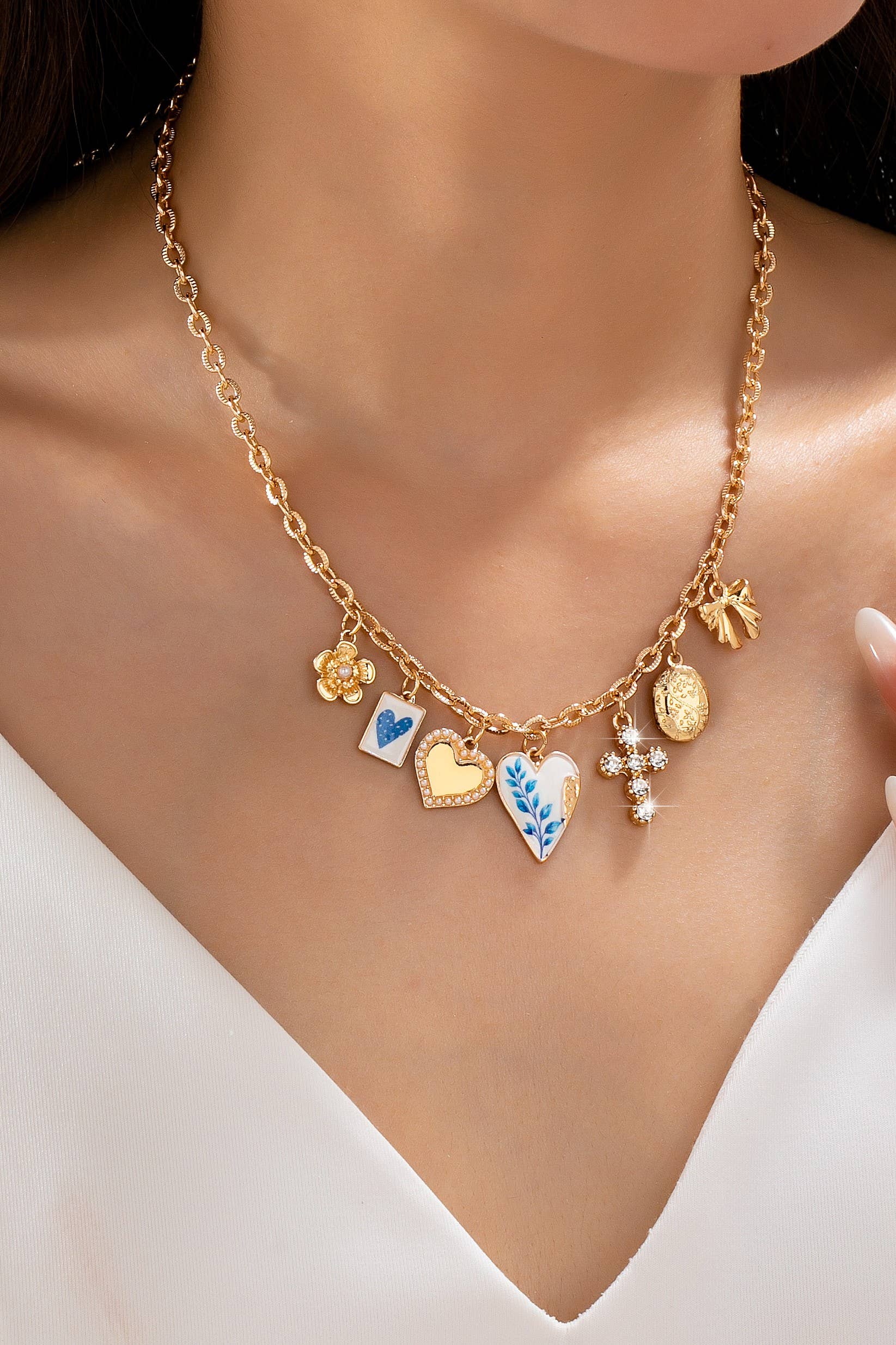 Wearing a white v-neck top, the person showcases the JOSSLYN by wall to wall Blue Vintage Coquette Charm Necklace, a gold chain adorned with six unique charms—hearts, a flower, a blue leaf design, a coin, a bow, and a cross.