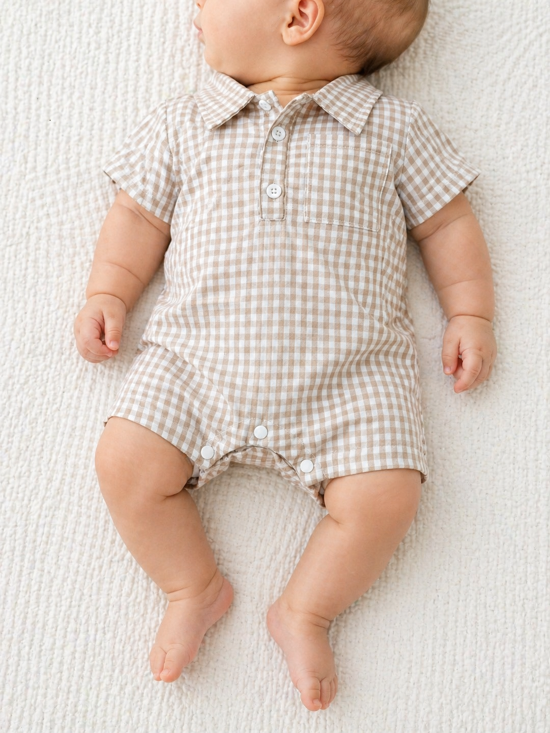 A baby in a Tiny Tot Bundles Beige Gingham Collard Romper with short sleeves lies on a textured white blanket, facing slightly to the side.