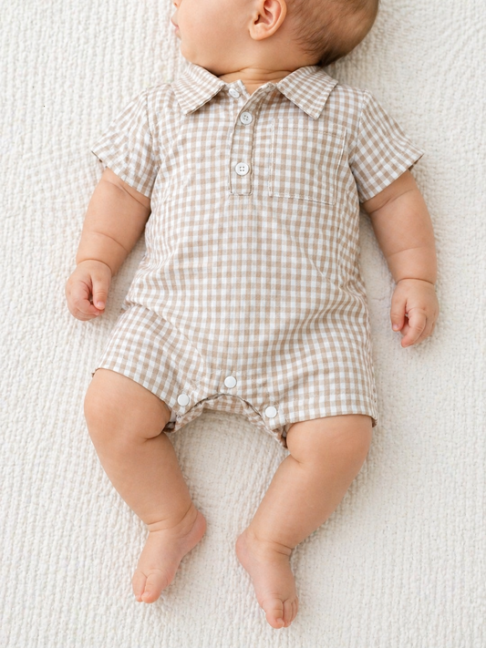 A baby in a Tiny Tot Bundles Beige Gingham Collard Romper with short sleeves lies on a textured white blanket, facing slightly to the side.