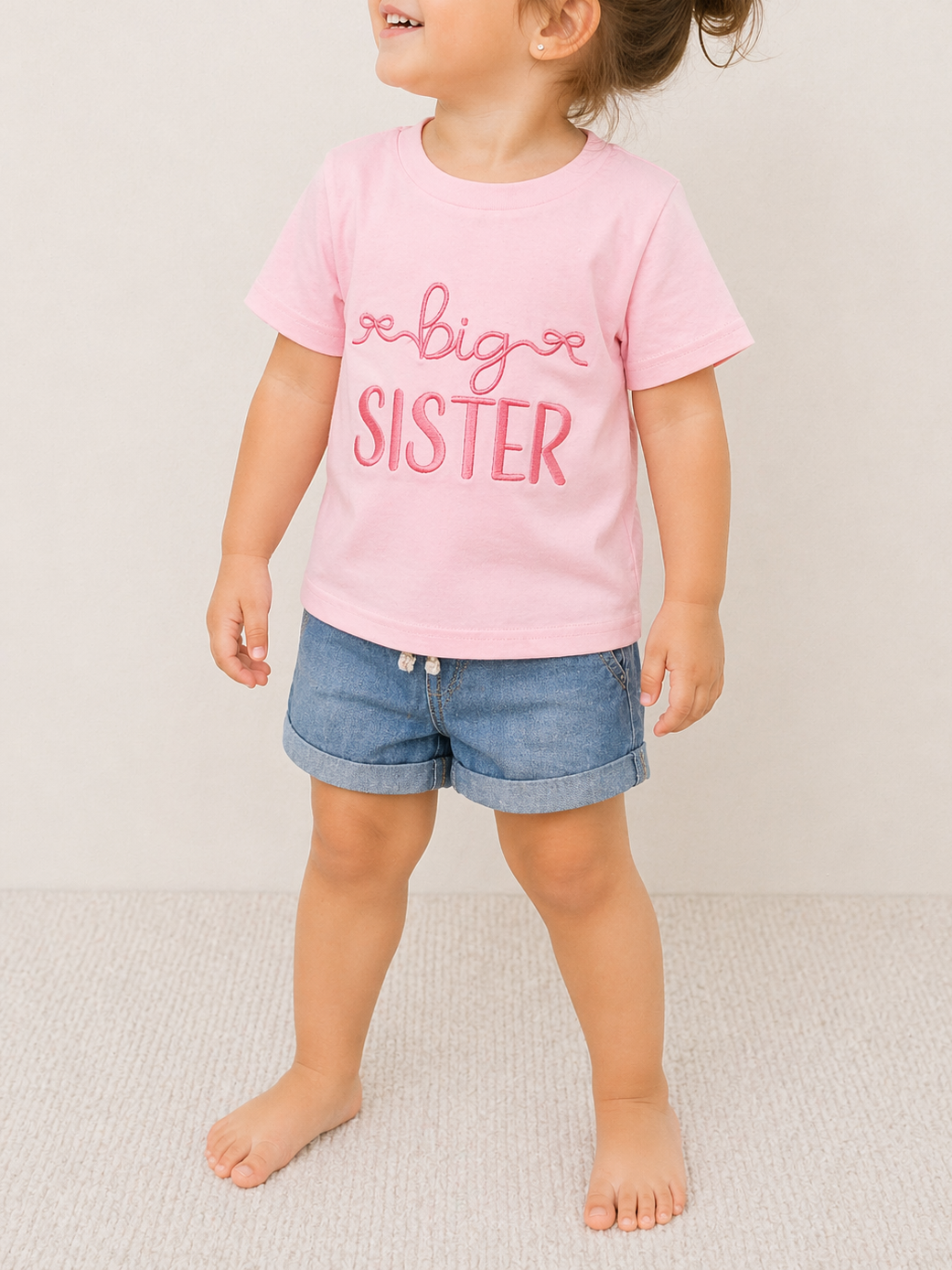 A young child stands barefoot on a light carpet, wearing the Tiny Tot Bundles Big Sister Bow T-Shirt and blue denim shorts. The child's face is partially out of the frame.