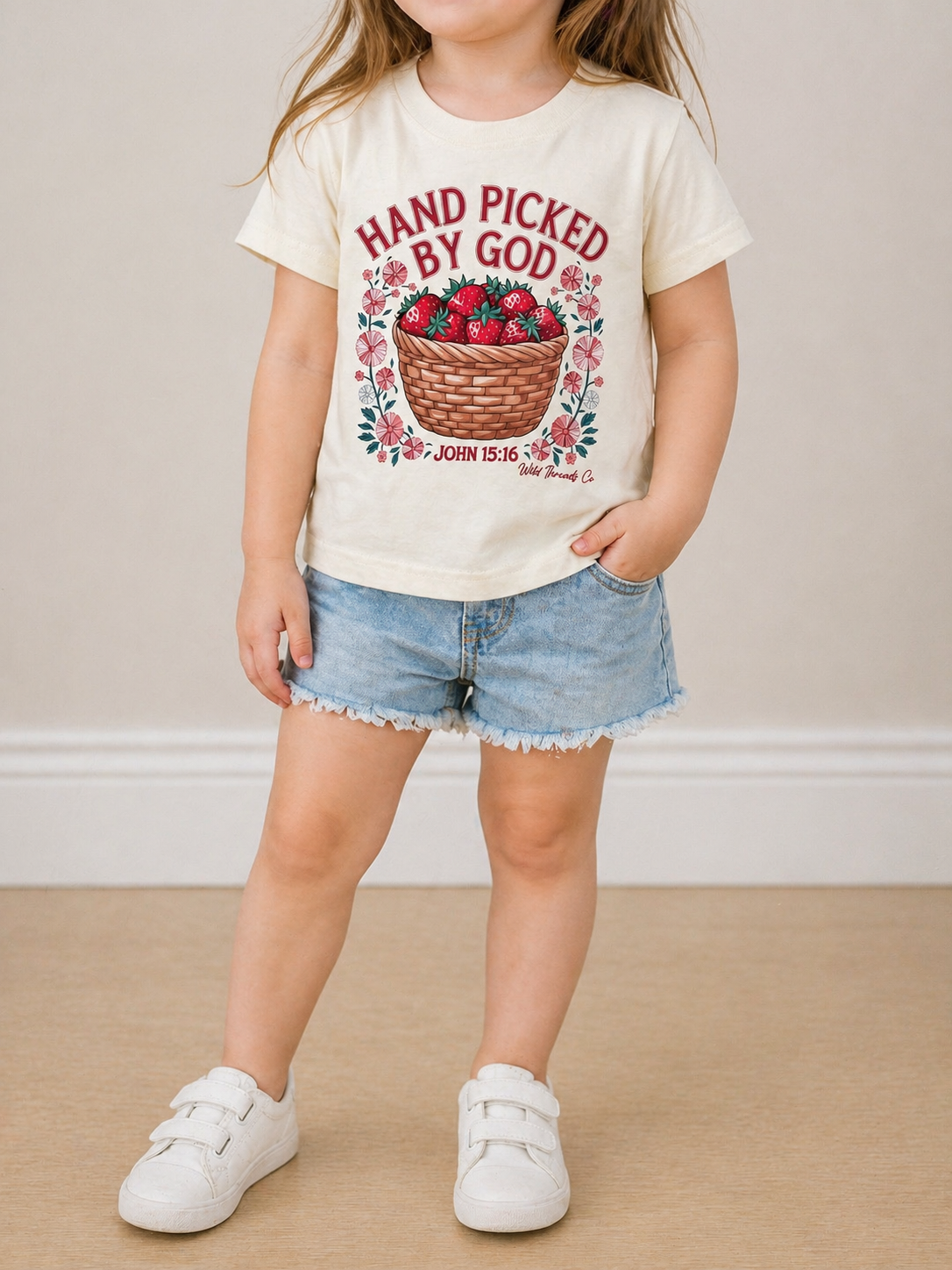 A young child indoors wears white sneakers, denim shorts, and the Wild Threads Co Hand Picked By God T-Shirt, featuring a basket of red flowers and “John 15:16” printed on it.