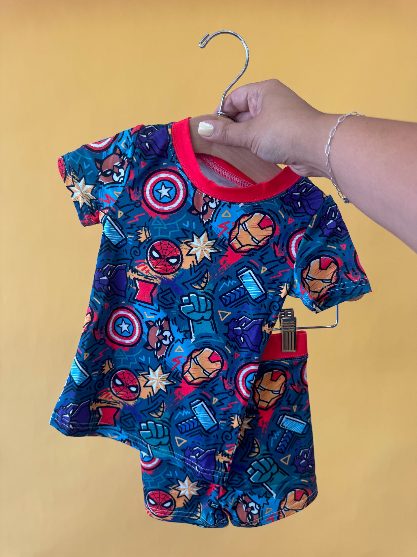 A hand holds a hanger displaying the Tiny Tot Bundles Hero Headquarters Lounge Set—a colorful, short-sleeve toddler outfit adorned with comic-style superhero symbols on a yellow background.