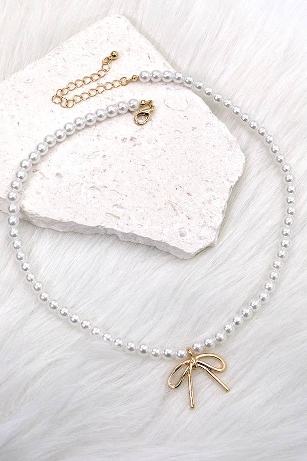 The JOSSLYN by wall to wall Pearl Chain Bow Charm Necklace, featuring lustrous pearls with a gold bow charm and an adjustable clasp, is displayed atop a white stone on a soft, white fur surface.