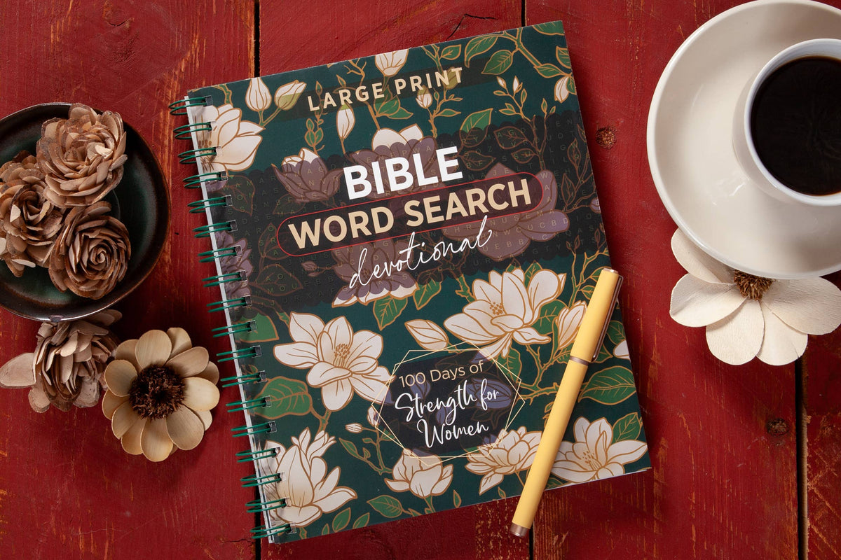 The “100 Days of Strength for Women Word Search Book” by BroadStreet Publishing Group, LLC, featuring a floral cover, is arranged alongside a pen, a cup of coffee, and decorative flowers on a red wooden table.