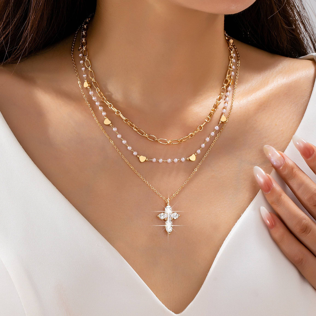 A woman styles her white v-neck top with the JOSSLYN by wall to wall Multi-Strand Pearl & Crystal Cross Necklace, featuring layered strands adorned with heart charms, pink beads, and a sparkling cross pendant.