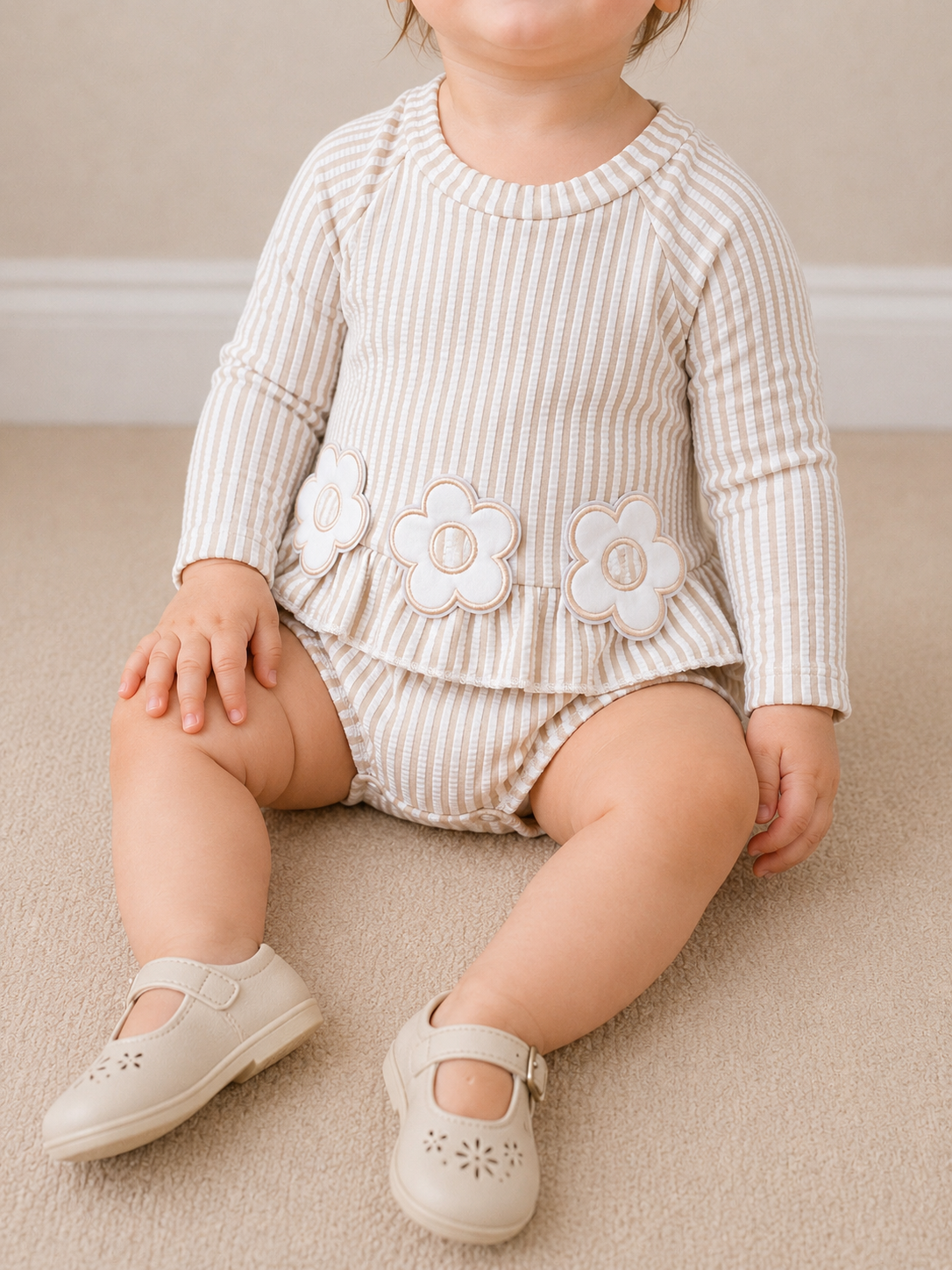 A young child sits on a beige carpet, wearing the Tiny Tot Bundles Petal Lane Swimsuit. The cream and tan striped suit features flower appliqués at the waist, paired with beige Mary Jane shoes. The child's face is partially out of frame.